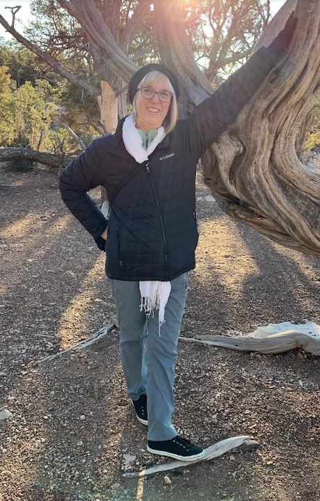 An older woman with glasses, wearing a black hat, black jacket, light-colored scarf, and light blue pants, standing outdoors beside a large, twisted tree, smiling at the camera with sunlight shining through the trees.