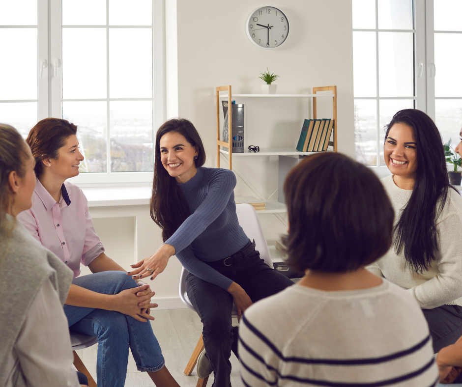 Group of five women sitting in a circle and talking in a bright room with large windows, a white wall, and a clock.