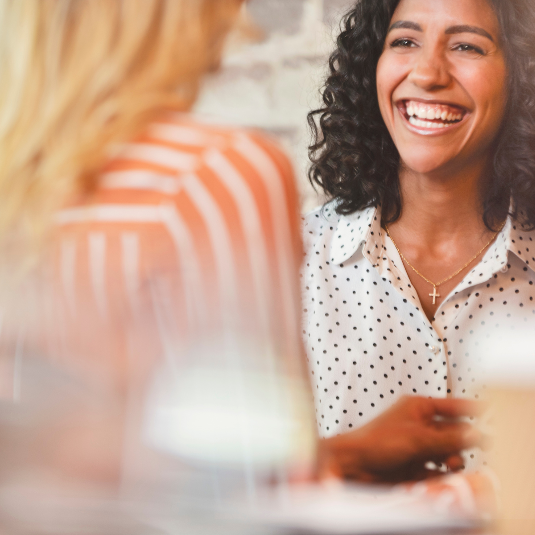 Two women with long hair, one with black curly hair wearing a white blouse with black polka dots, the other with blonde hair wearing a red and white striped top, laughing and having a conversation in a casual setting.