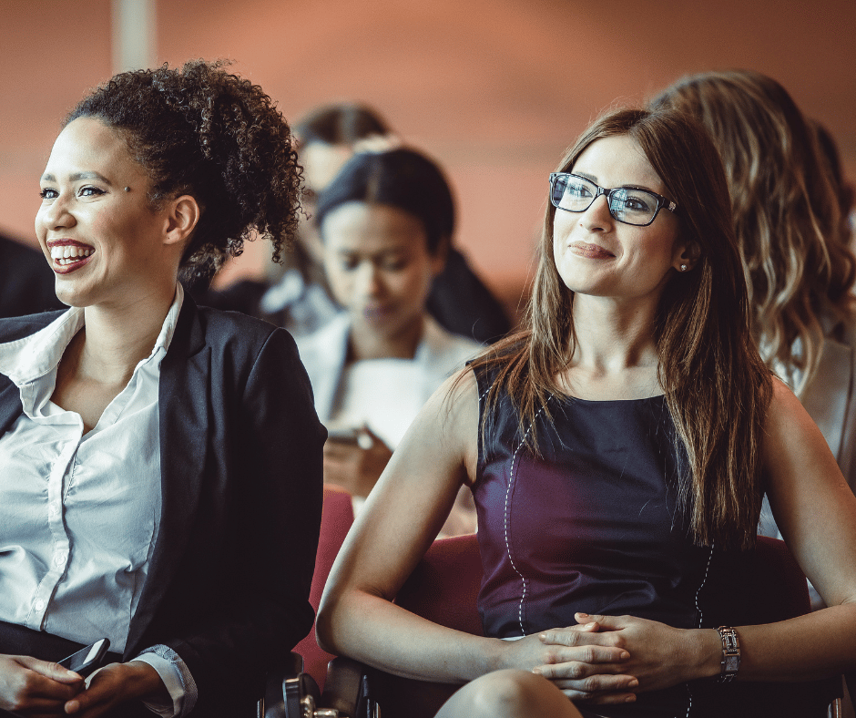 Two women sitting and smiling at a conference or seminar, with other attendees in the background.