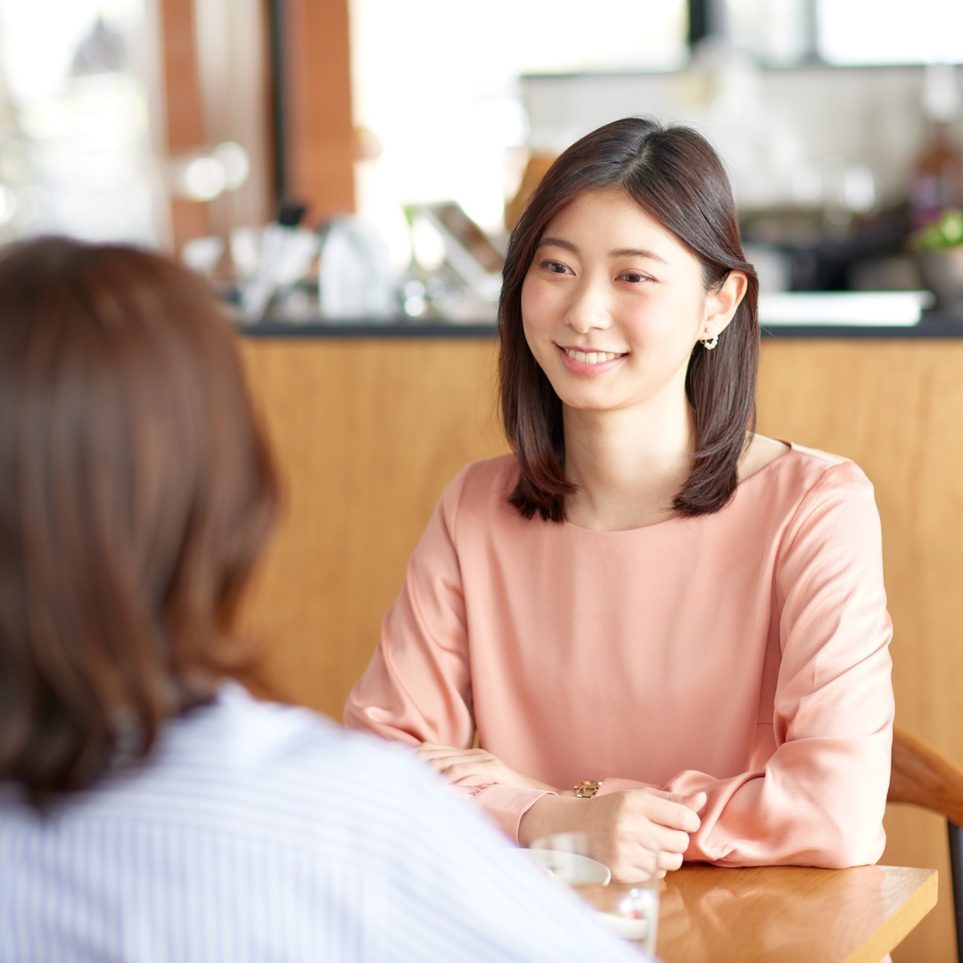 A woman with shoulder-length dark brown hair wearing a pink satin blouse, smiling and engaging in conversation with another person at a wooden table in a bright, modern cafe.