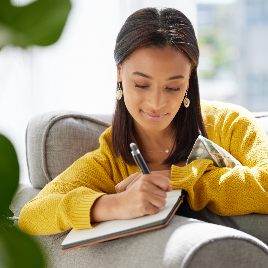 A woman wearing a yellow sweater sitting on a sofa, writing in a notebook with a pen, near a window with sunlight coming through.