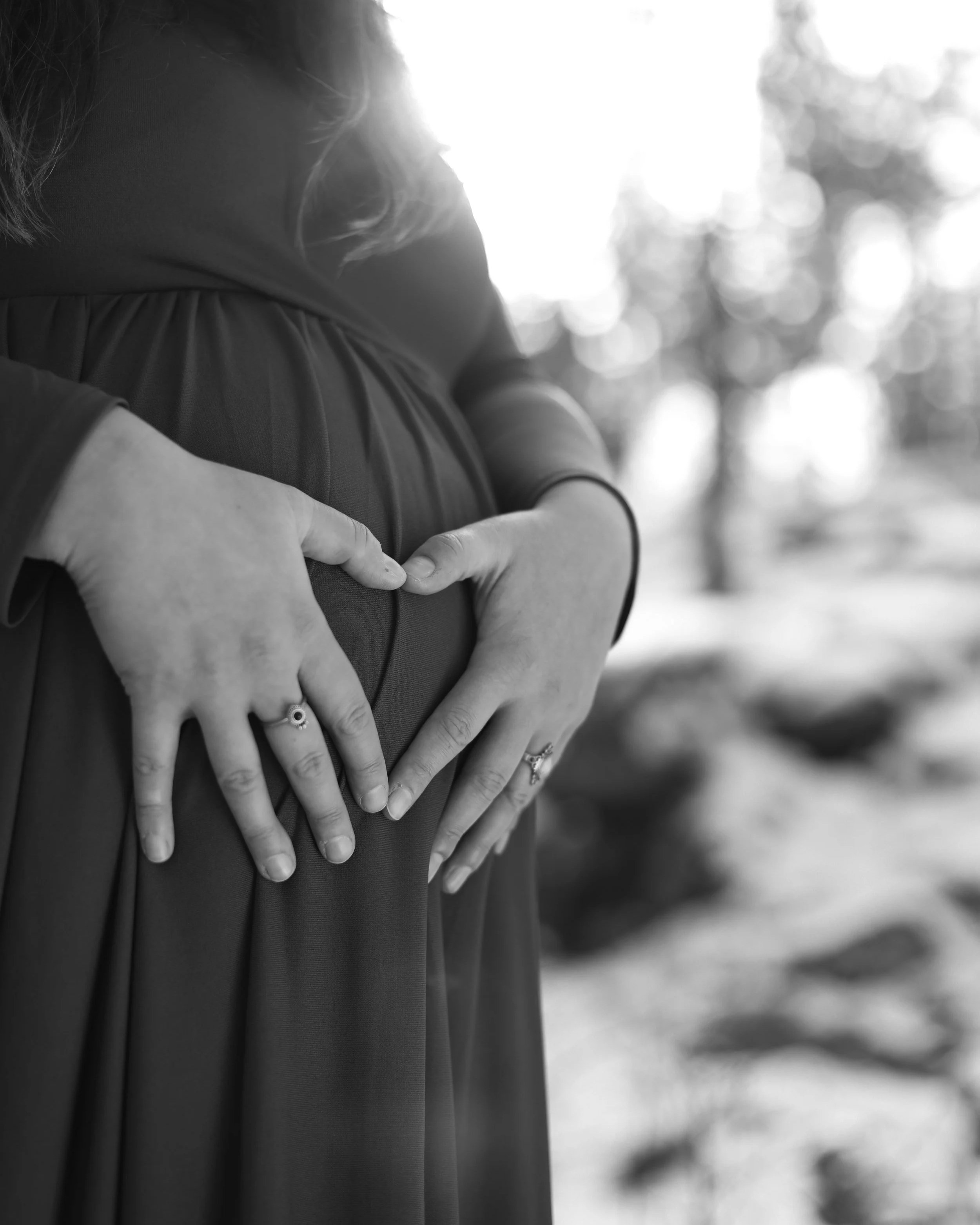 Two people making a heart shape with their hands on a person's belly, suggesting pregnancy; black-and-white photo with blurred trees in the background.