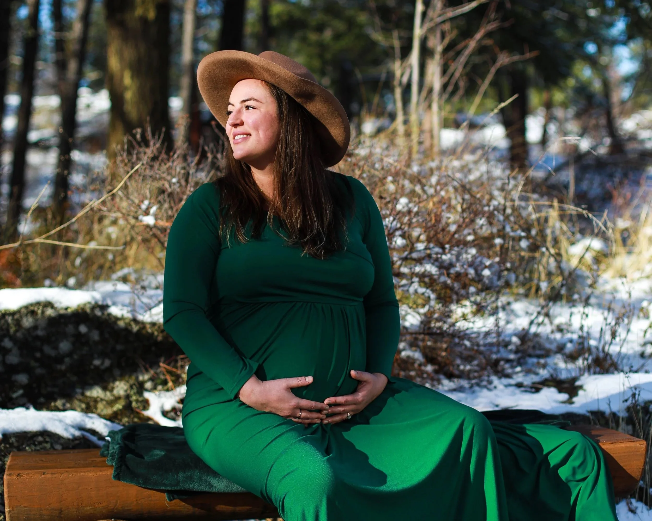 A pregnant woman in a green dress and wide-brimmed hat sitting on a wooden bench outdoors in a snowy forest, gently holding her belly.