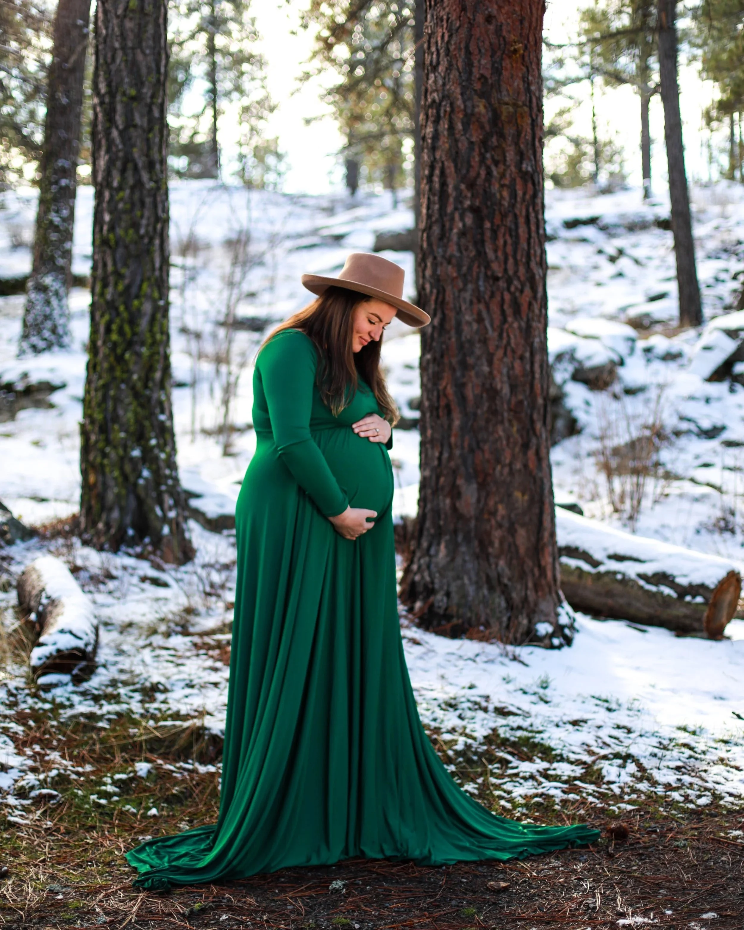 Pregnant woman in a green dress and tan hat standing in a snowy forest, gently holding her belly and smiling.