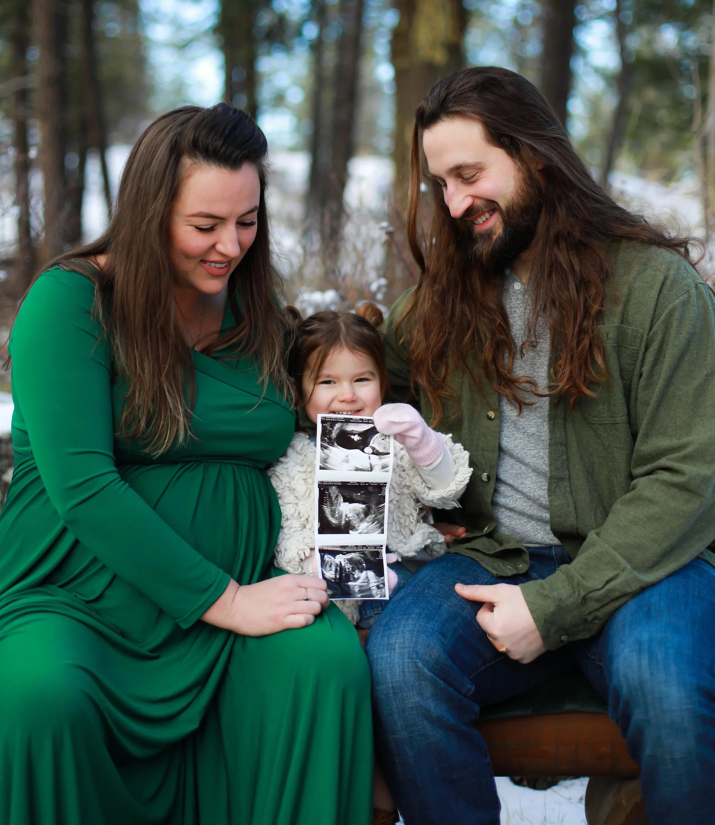 A pregnant woman in a green dress, holding ultrasound images, sitting outdoors with a man and a young girl in a snowy forest setting, all smiling.