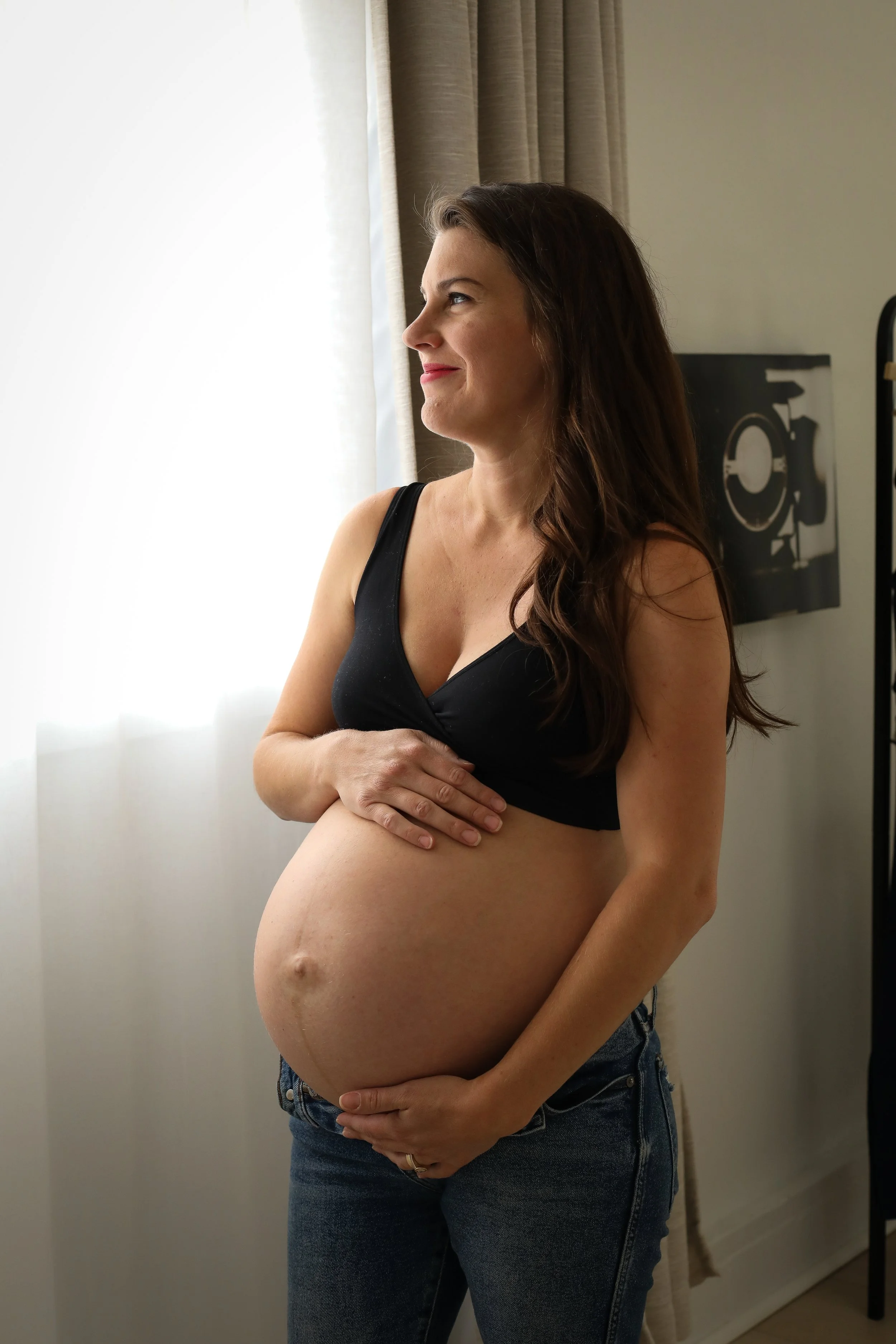 Pregnant woman standing indoors near a window with curtains, gently holding her belly with both hands, wearing a black top and jeans.