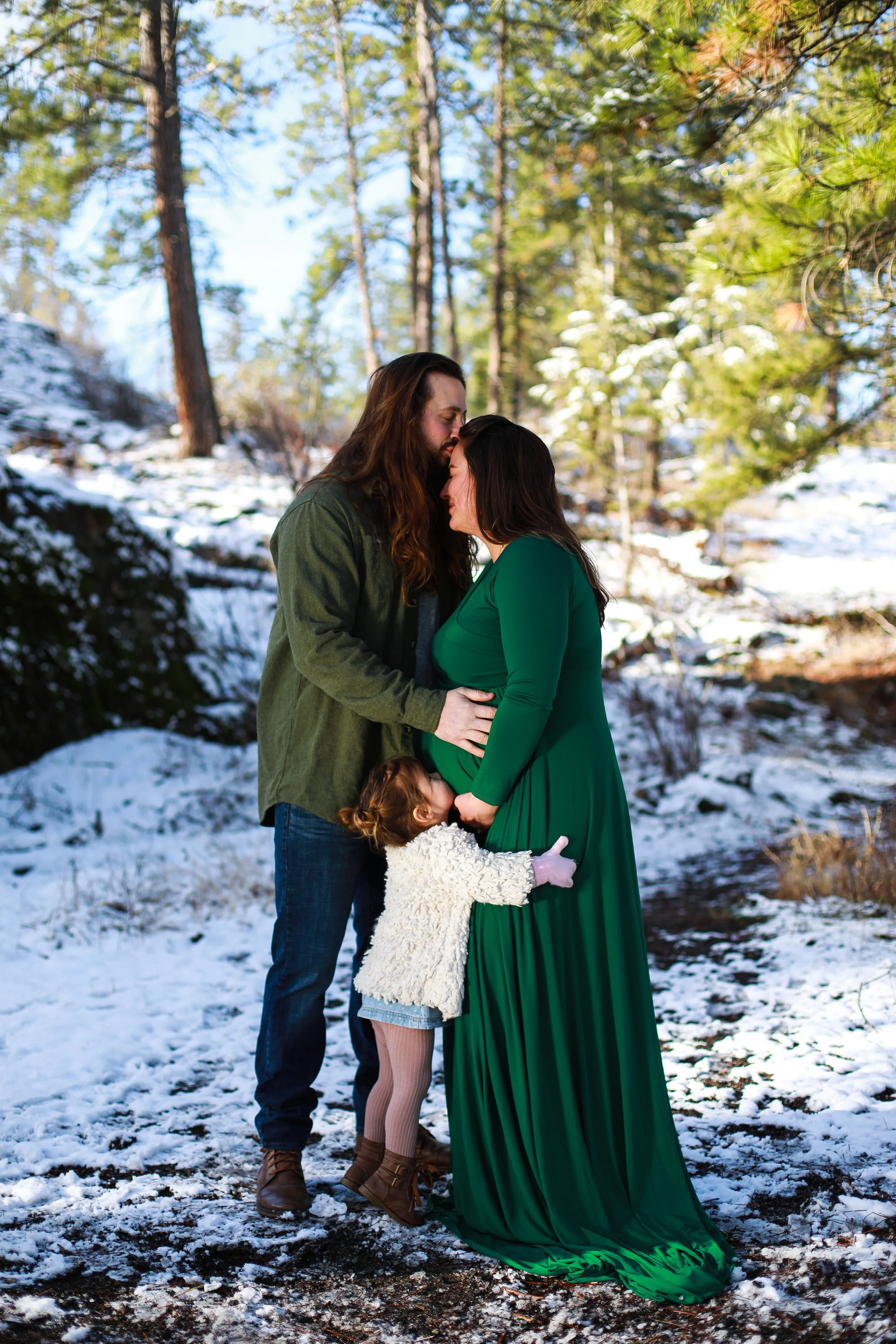 A family of three, including a pregnant woman in a green dress, a man with long hair, and a young girl in a white sweater, standing closely together outdoors amid snow-covered ground and tall pine trees.
