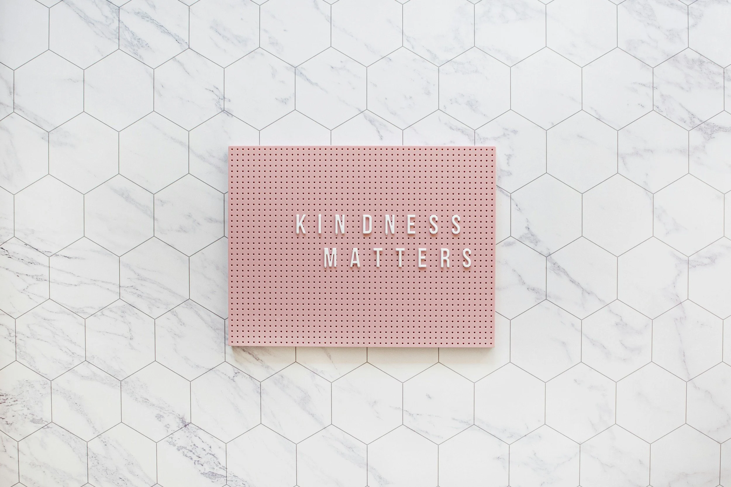 Pink board with white letters spelling "KINDNESS MATTERS" on white marble hexagon tiled floor.