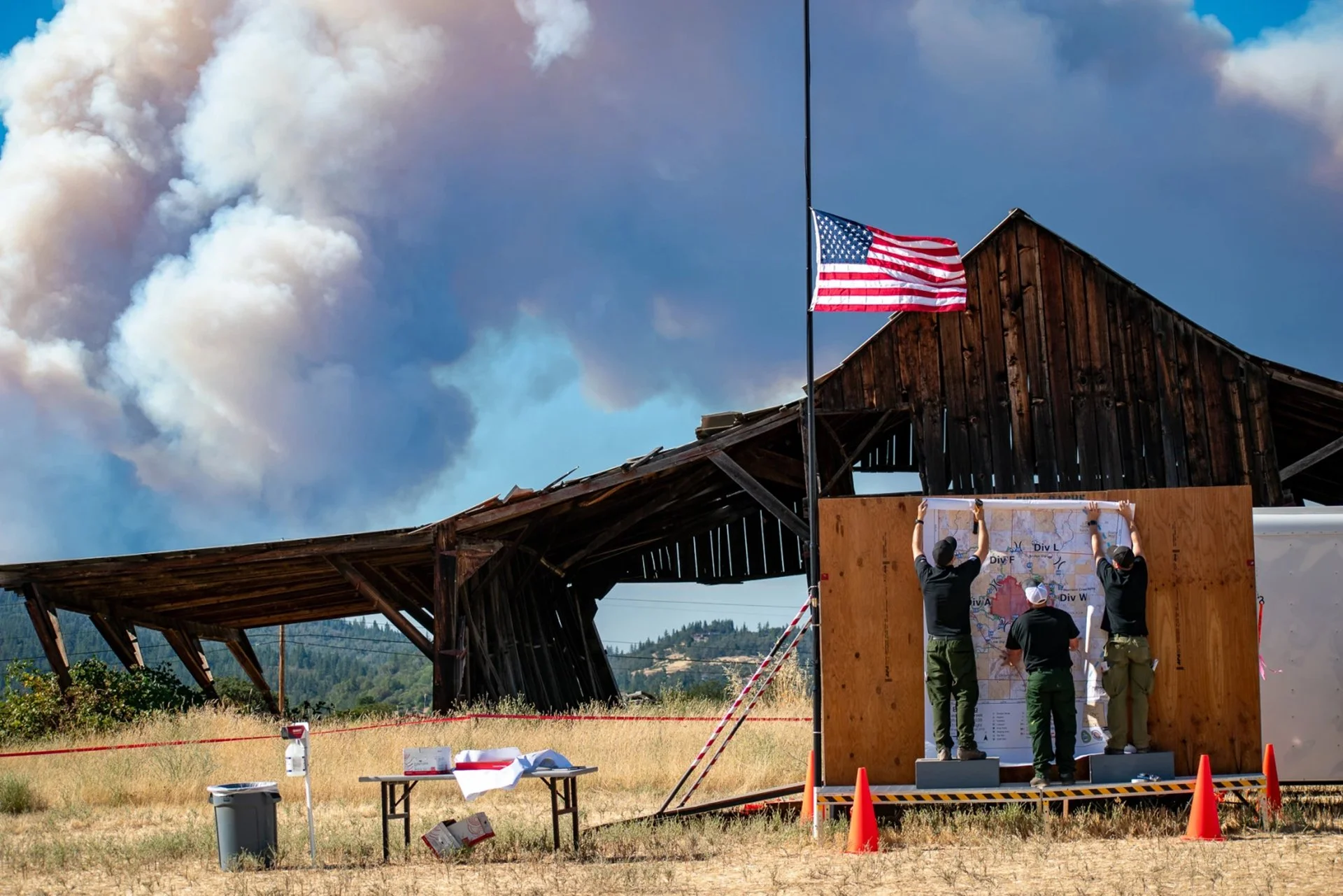 People hanging a map on a wooden wall with an American flag on a pole, under a partly cloudy sky with smoke or clouds in the background at an outdoor event.
