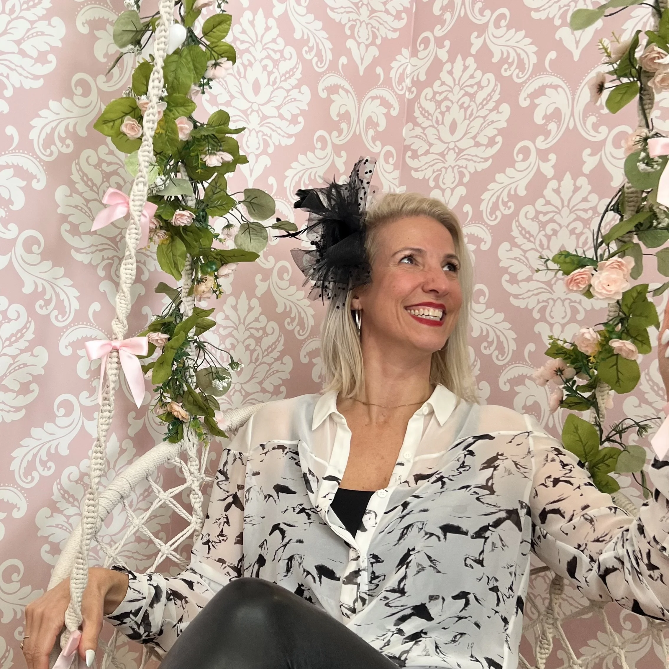 Smiling woman with blonde hair wearing a white blouse with black animal print, black shirt underneath, and a black floral fascinator, sitting on a white woven swing decorated with pink roses and green leaves against a pink and white damask patterned wall.