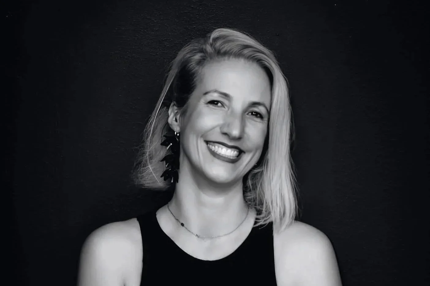 Black and white portrait of a smiling woman with blonde, shoulder-length hair, wearing a sleeveless top, large earrings, and a delicate necklace, against a dark background.