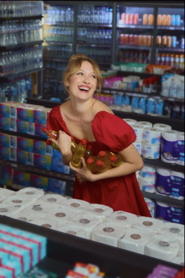 A woman in a red dress holding bottled drinks and laughing inside a store aisle with shelves of grocery items in the background.