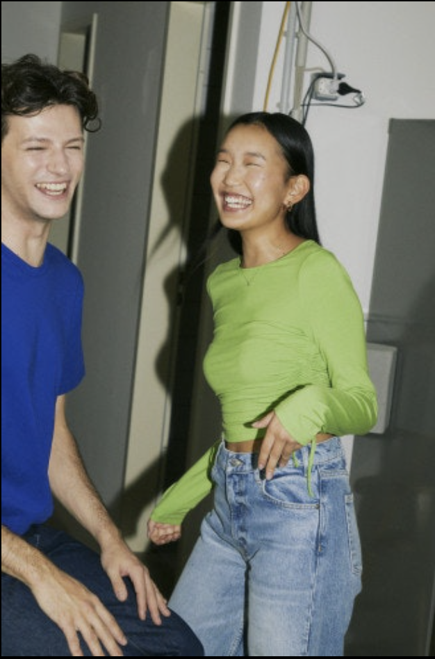 Two young adults, a man and a woman, are laughing and enjoying a moment together indoors. The man has curly dark hair and is wearing a blue shirt, while the woman has straight black hair and is wearing a lime green long-sleeve top and high-waisted jeans.