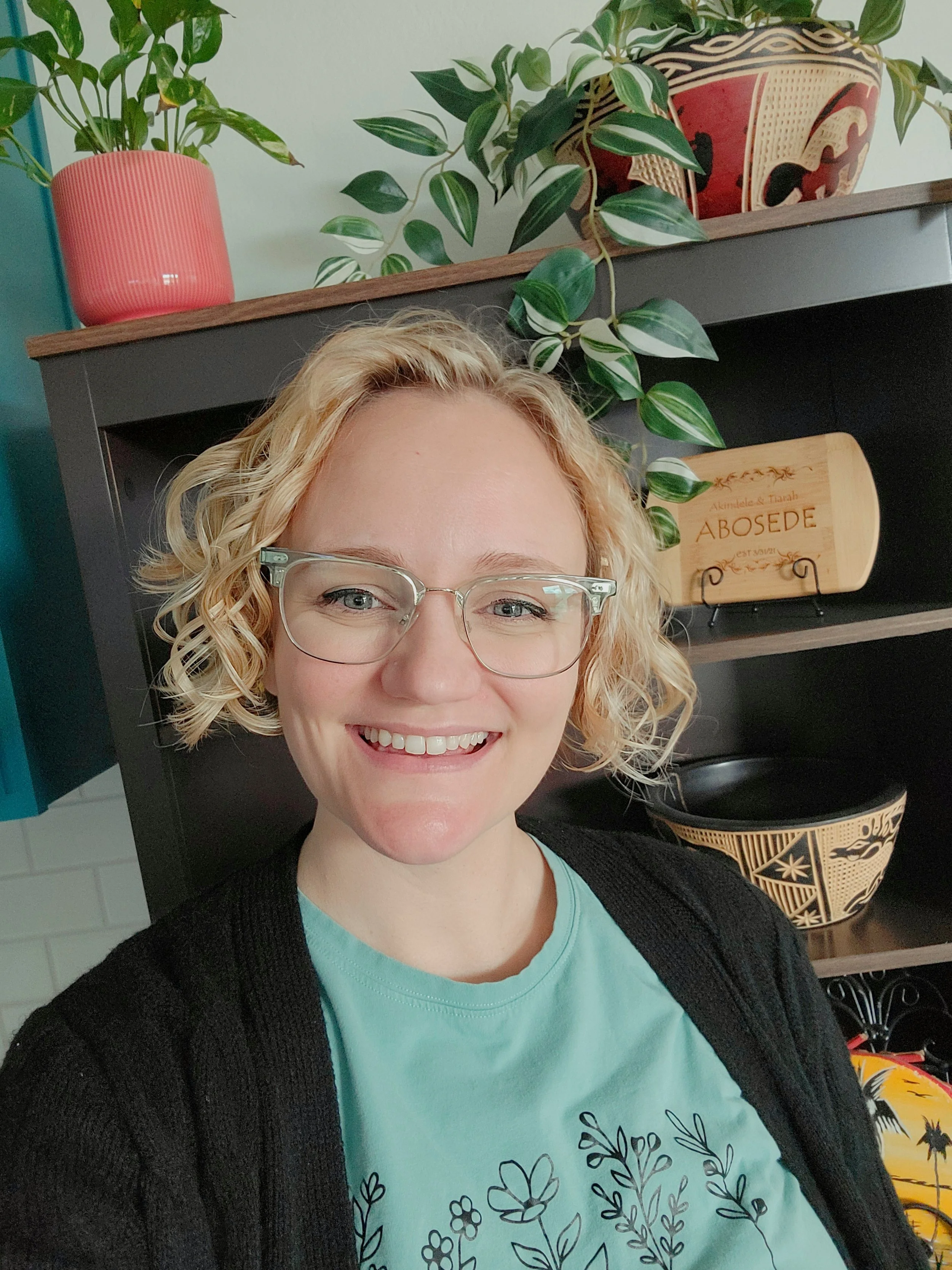 A woman with curly blonde hair and glasses smiling in front of a black shelf with decorative items and plants.