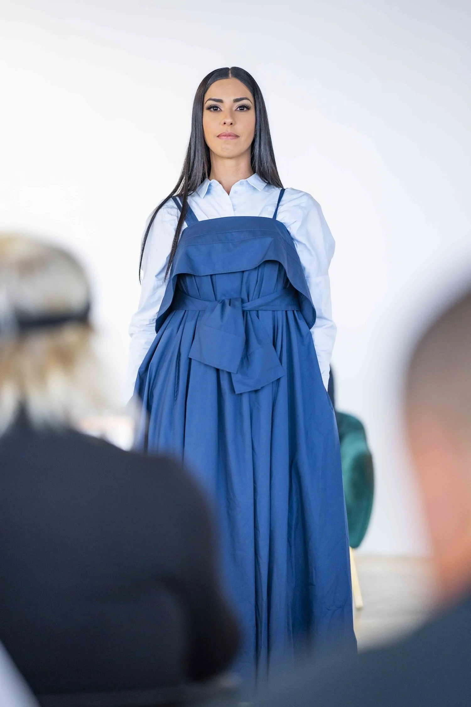 Woman with long dark hair wearing a white shirt, a blue dress layered over, and sneakers, walking toward the camera against a white background.