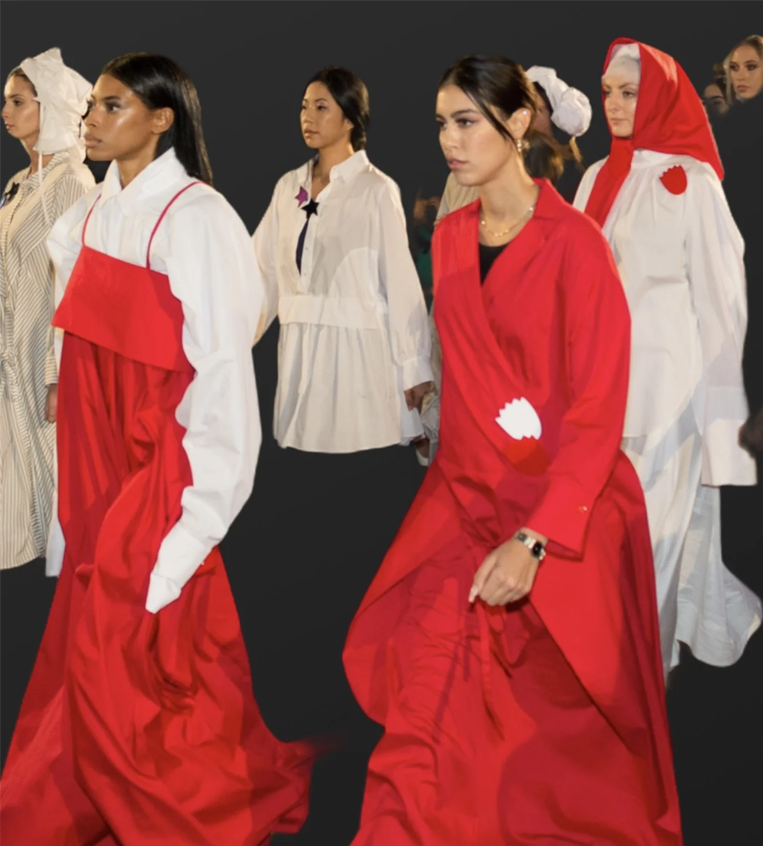 Multiple models walking on a runway, dressed in fashion outfits with prominent red and white colors, featuring layered and structured designs, against a black background.