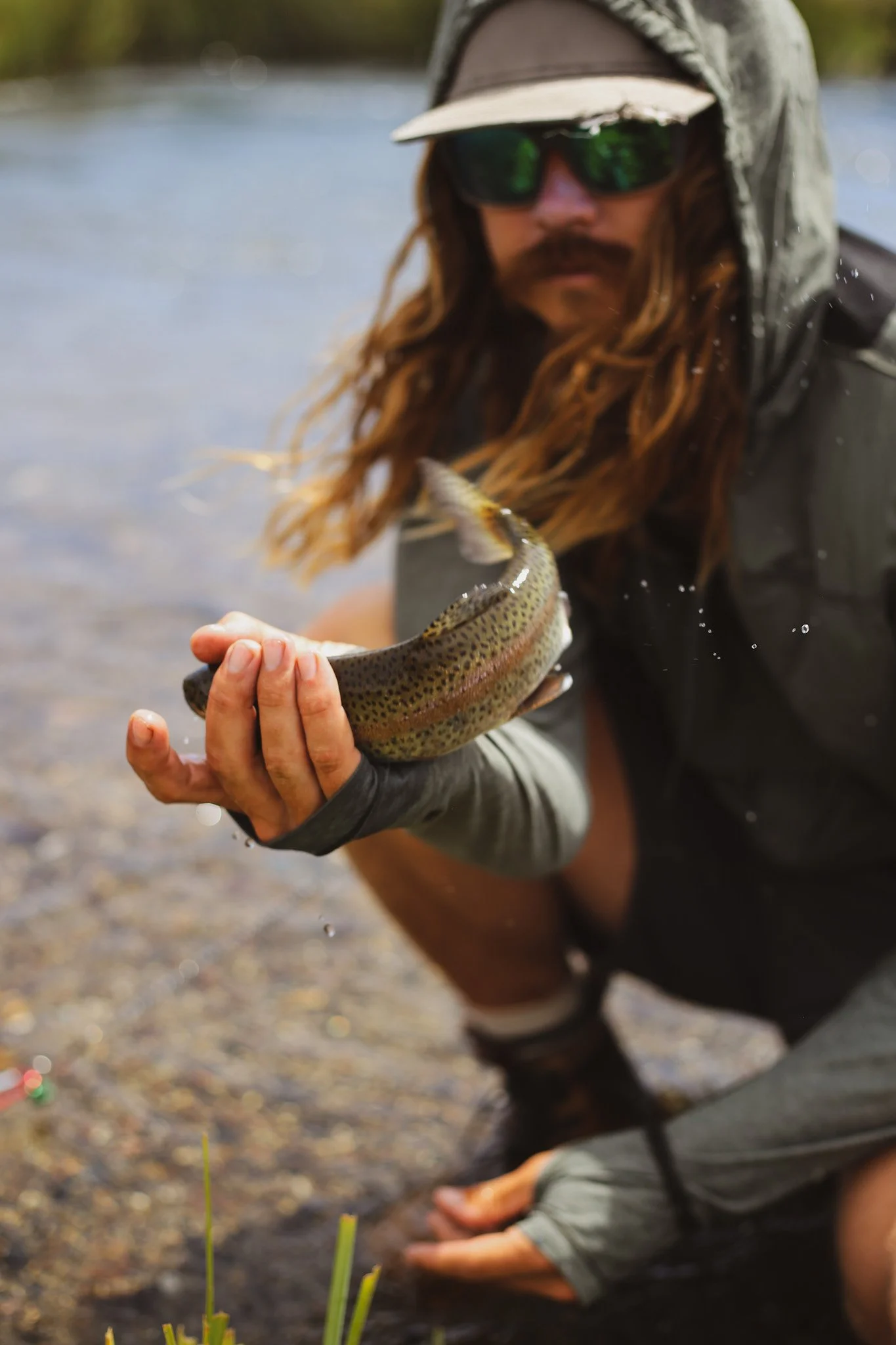 Man with long hair, sunglasses, and a hoodie holding a fish near the water's edge.