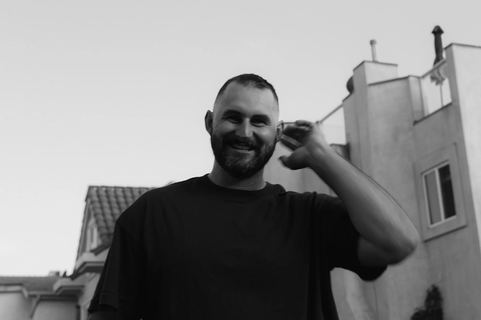 A smiling man with a beard talking on a mobile phone outdoors in front of houses.