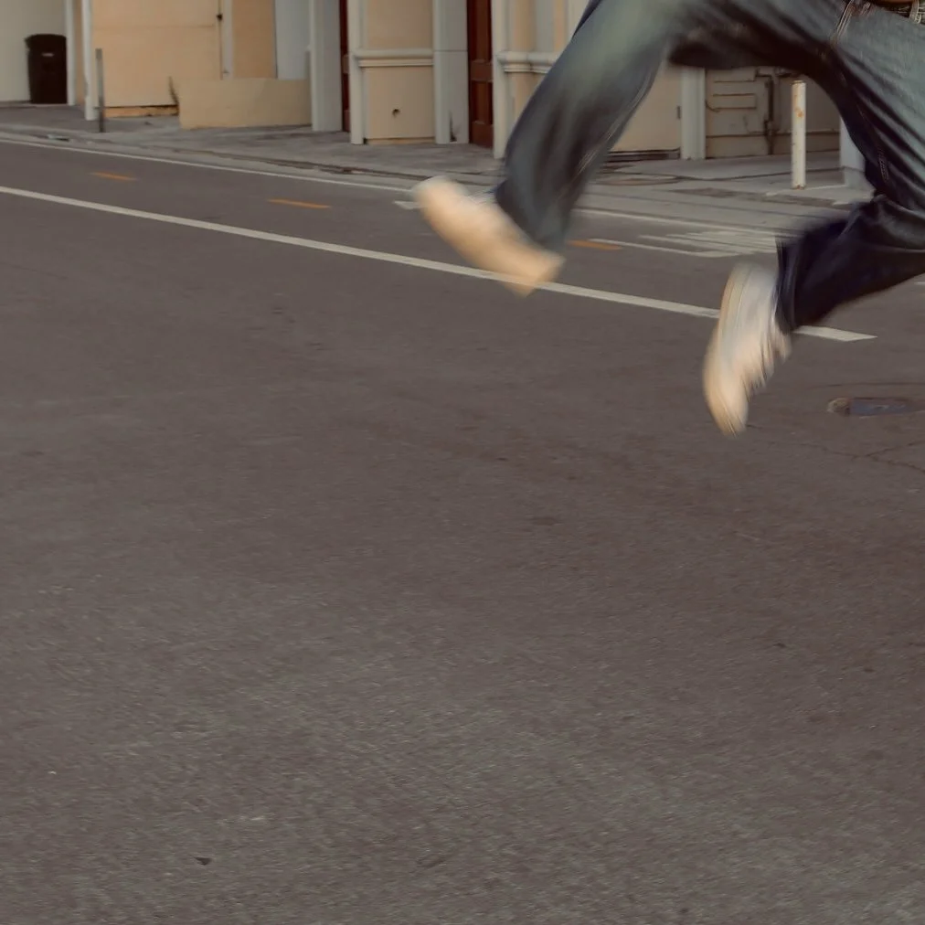 Person running or jumping on a city street wearing jeans and sneakers, with buildings and road markings visible in the background.