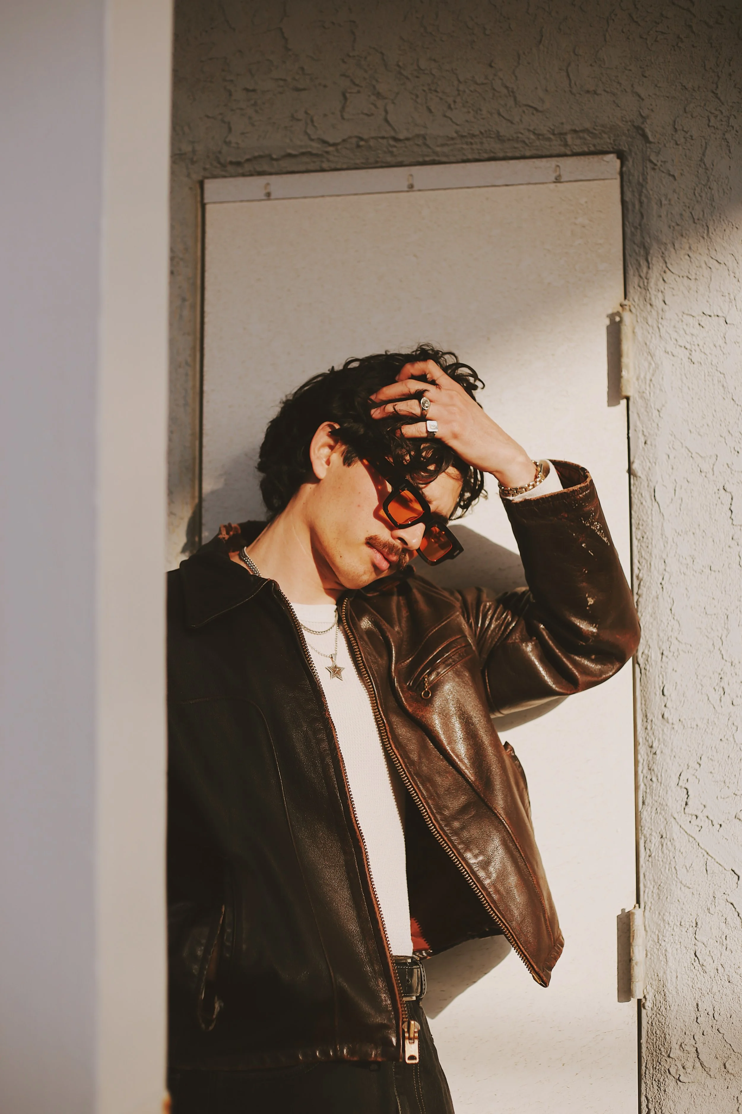 A man with dark hair, wearing sunglasses, a white shirt, and a brown leather jacket, is standing against a textured wall, touching his hair with one hand.