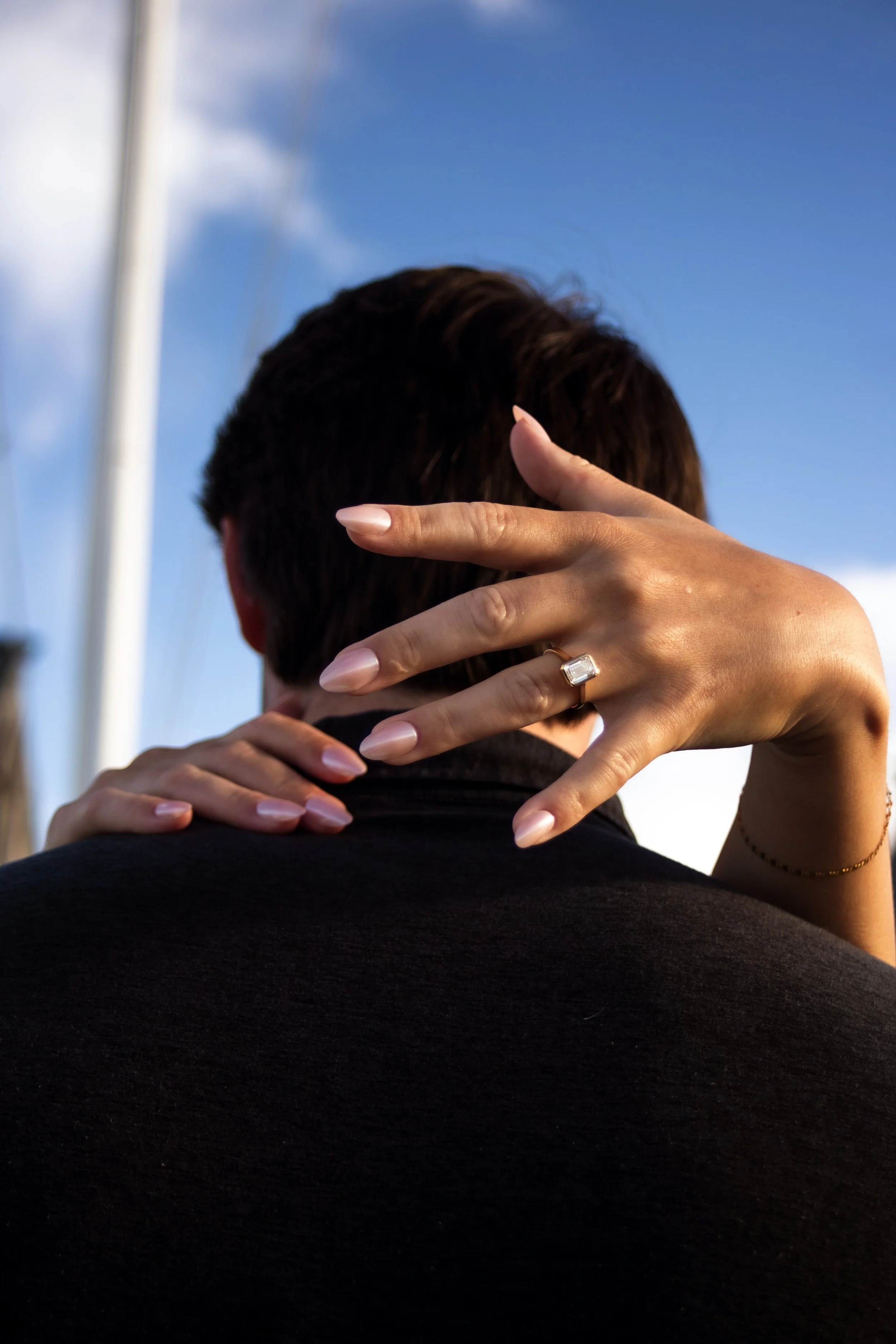 A woman with manicured nails and a large ring on her finger embraces a man whose face is not visible, outdoors against a blue sky with some clouds.