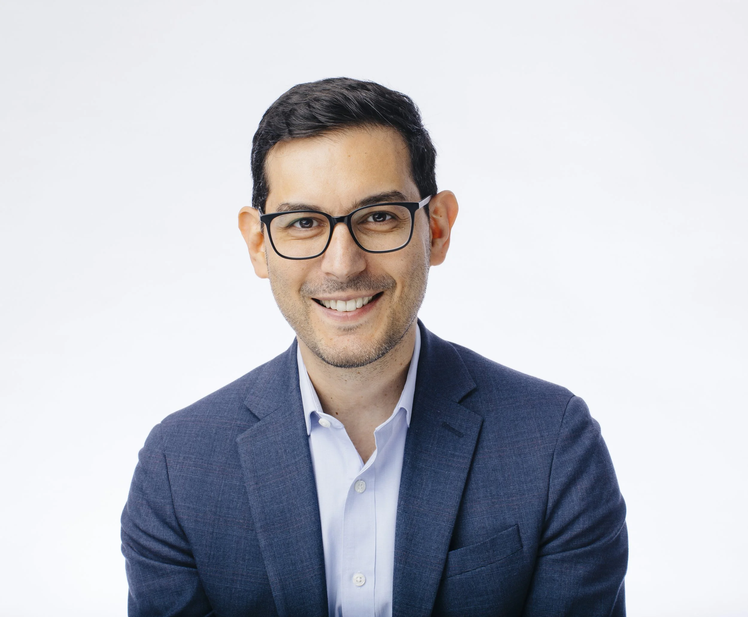 A portrait of a young man with dark hair, glasses, wearing a blue suit jacket and a light blue shirt, smiling against a plain white background.