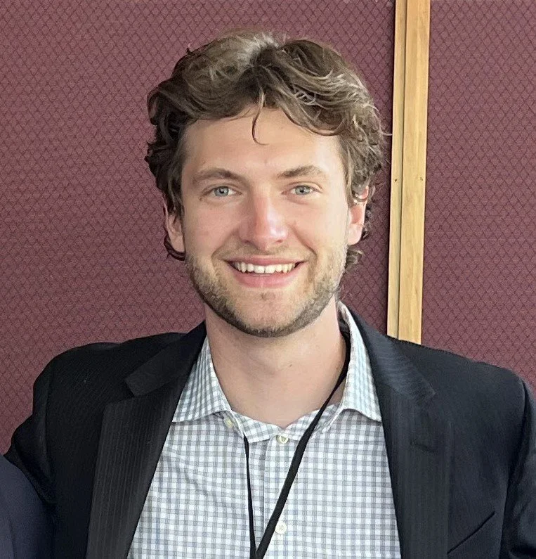 A young man with wavy light brown hair, fair skin, and a beard, smiling and wearing a black blazer over a checkered shirt, standing in front of a red textured wall.