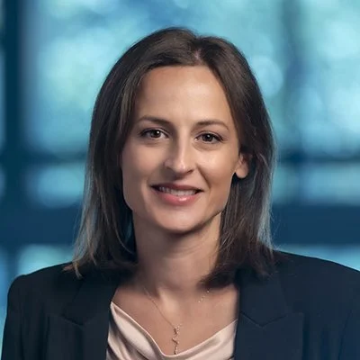 A woman with shoulder-length brown hair wearing a dark blazer and a light-colored top, smiling at the camera with a blurred blue and green background.