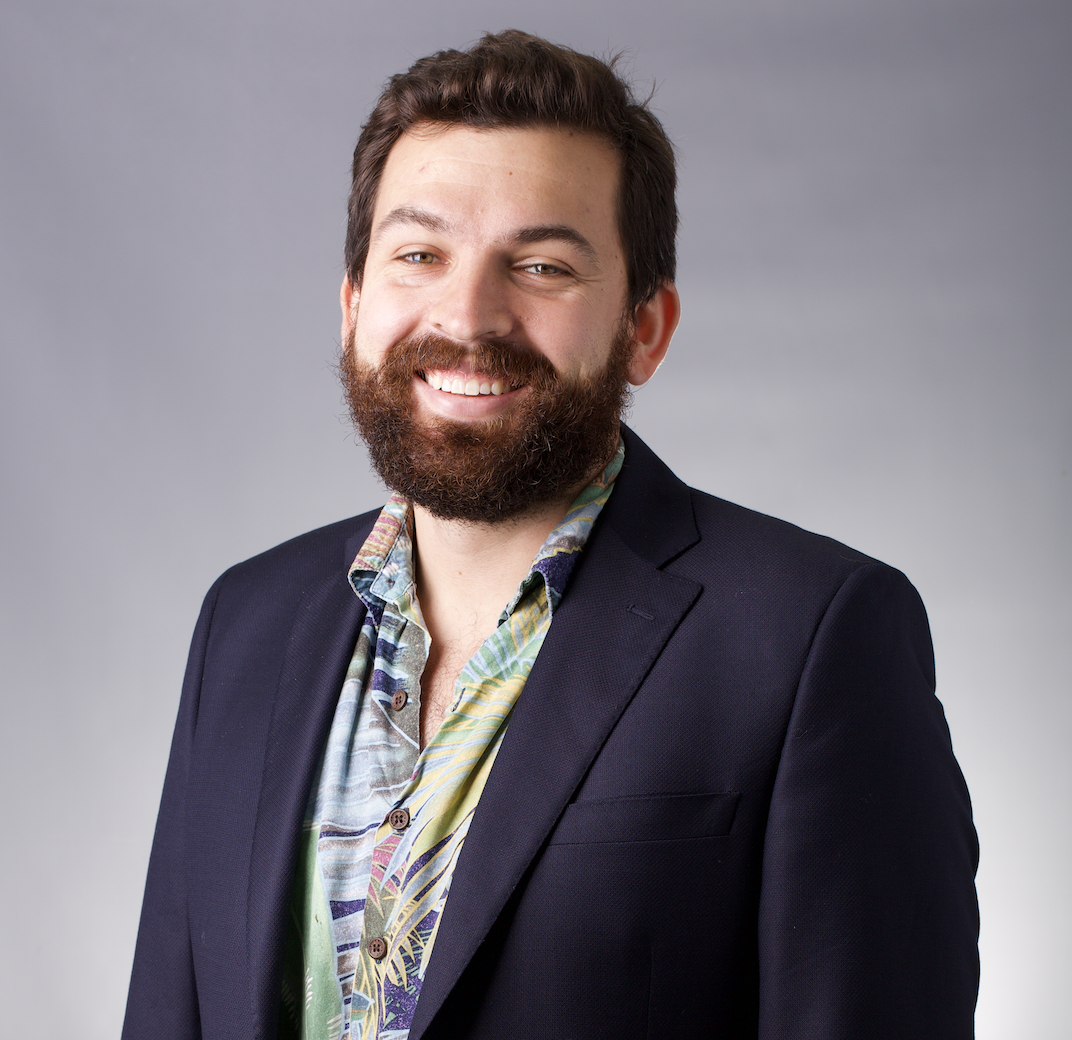 A man with a beard and mustache smiling, dressed in a colorful shirt and a black blazer against a plain gray background.