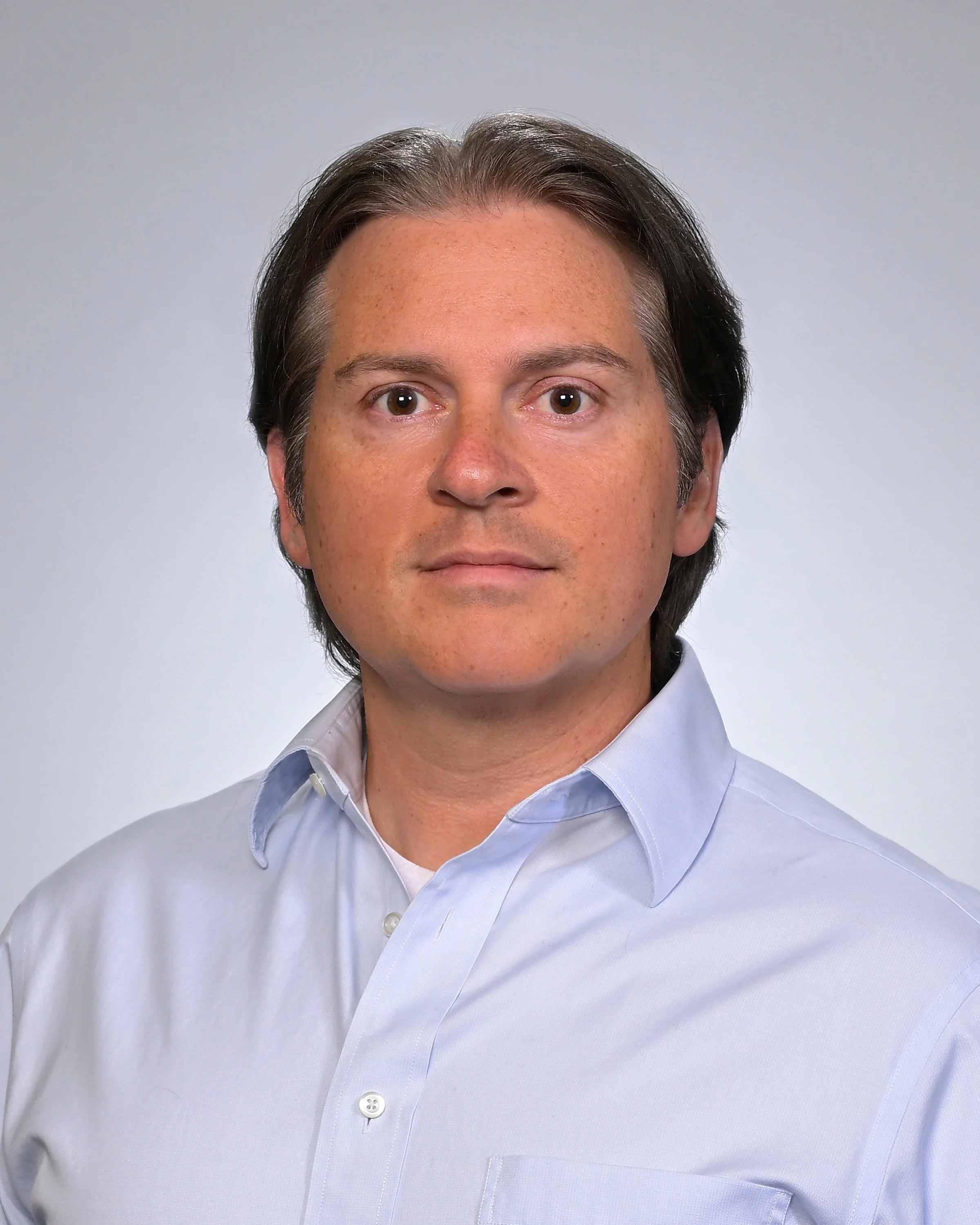 A man with dark hair and light skin wearing a light blue dress shirt, posing against a neutral background.
