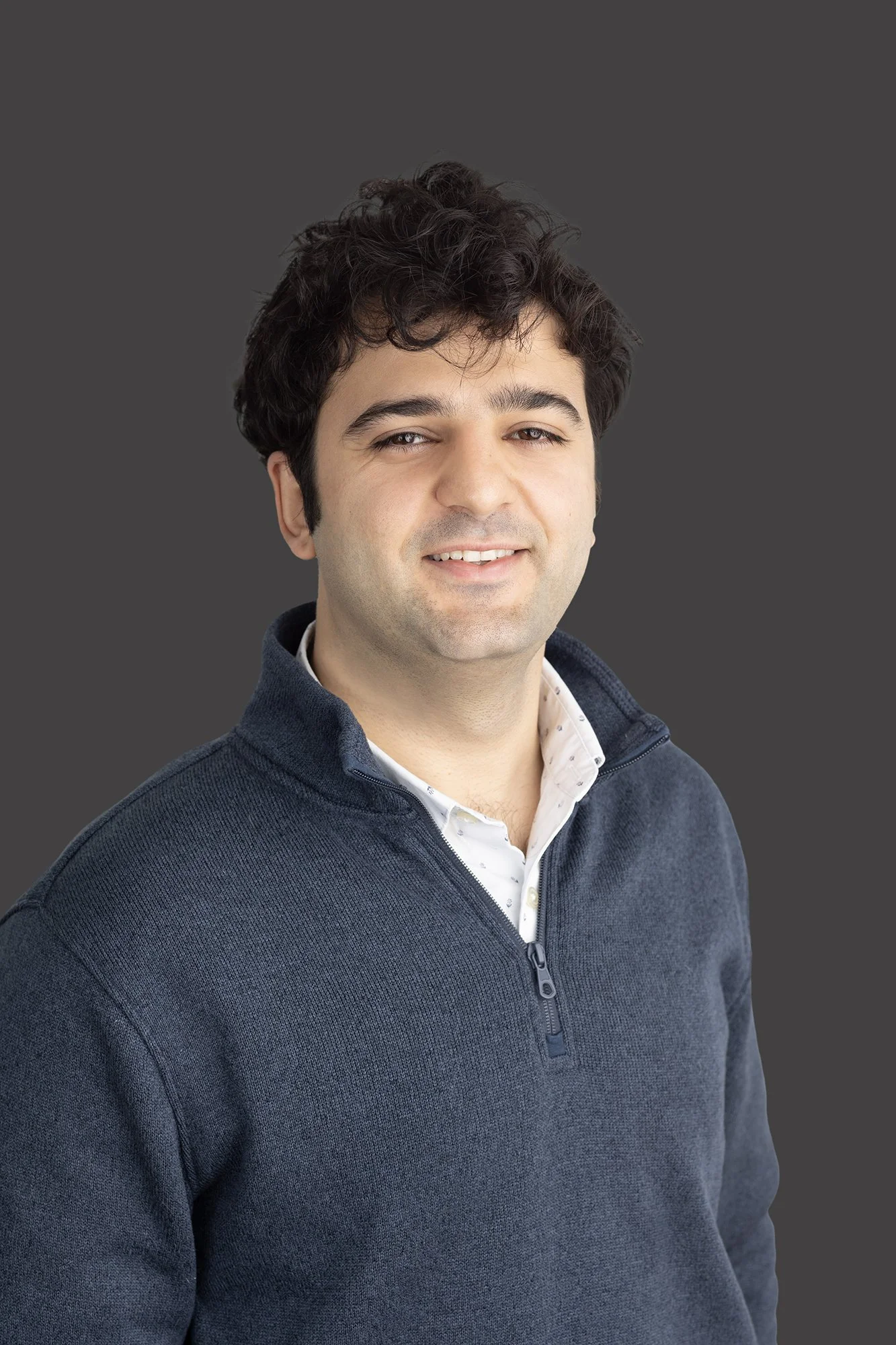 A young man with dark, curly hair, wearing a dark blue zip-up sweater over a collared shirt, smiling slightly against a plain gray background.