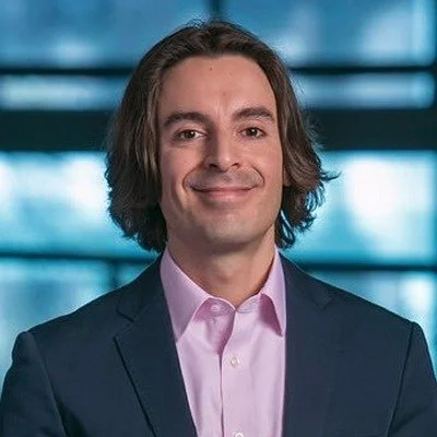 A man with shoulder-length brown hair wearing a dark suit and light pink shirt, standing in front of a blurred office background, smiling at the camera.