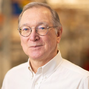 Portrait of an older man with glasses, wearing a white collared shirt, in an indoor setting with warm lighting.