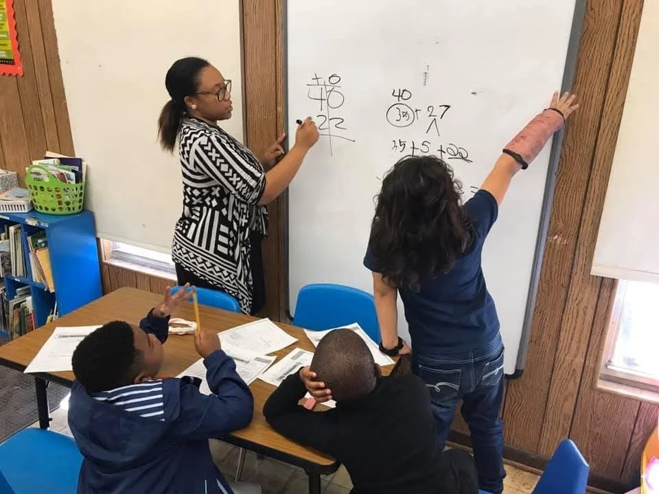 A teacher and three students in a classroom, with the teacher at the whiteboard writing math problems. One girl is reaching up to the whiteboard, and two boys are sitting at a table with open notebooks and pencils, paying attention to the lesson.