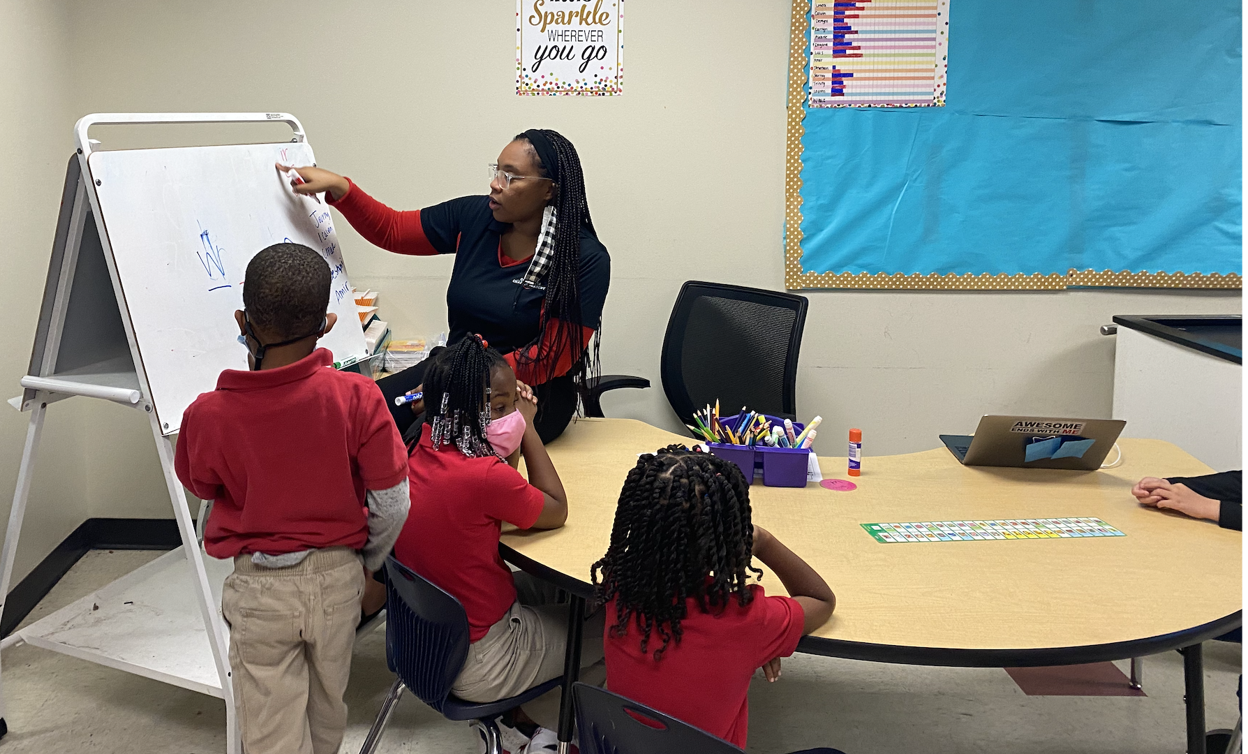 A teacher instructs young students in a classroom, standing by a whiteboard with writing, while children sit around a table, some wearing masks and one child standing in a red shirt.