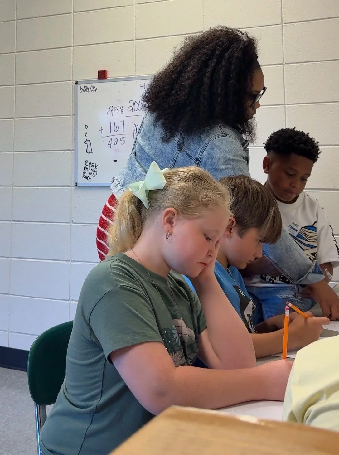 A teacher and three students in a classroom, with a whiteboard in the background showing math calculations.