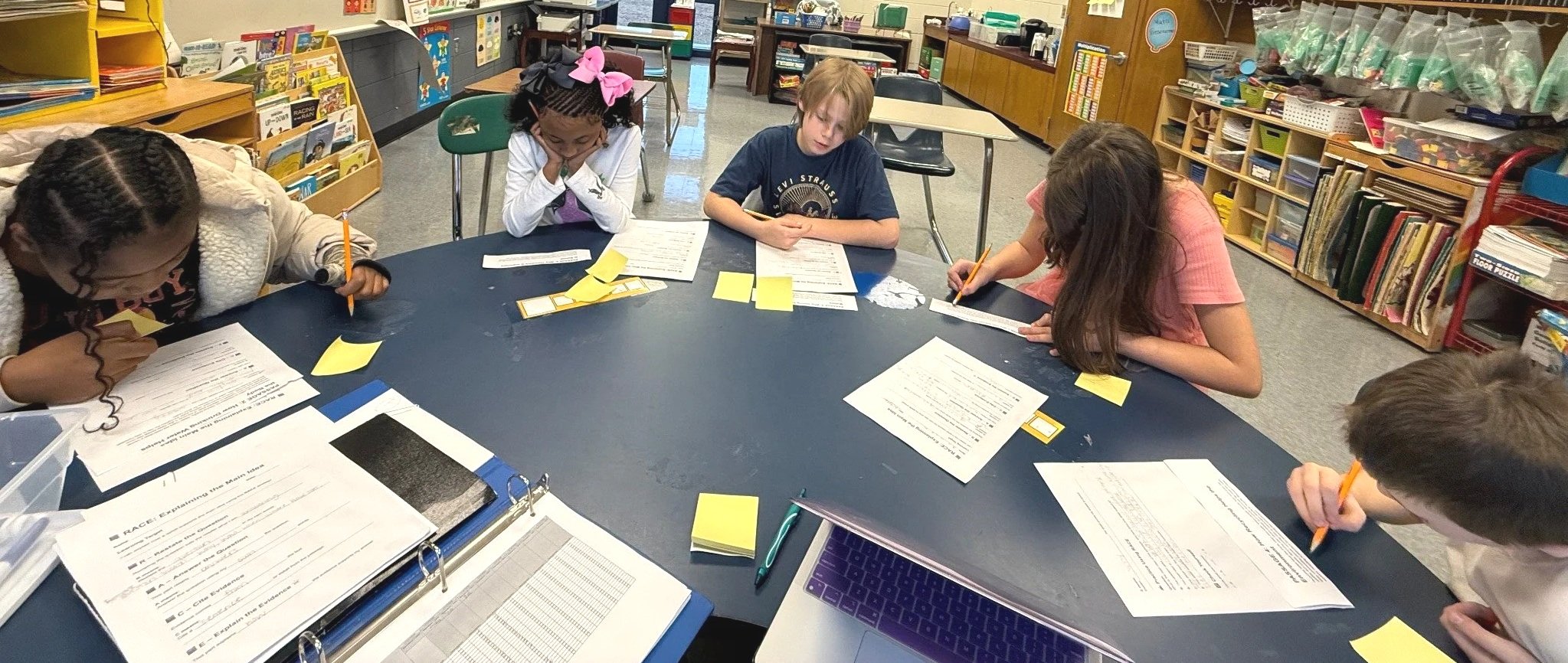 Four children sit around a blue table in a classroom, working on papers with pencils. The classroom has bookshelves filled with books and educational materials in the background.