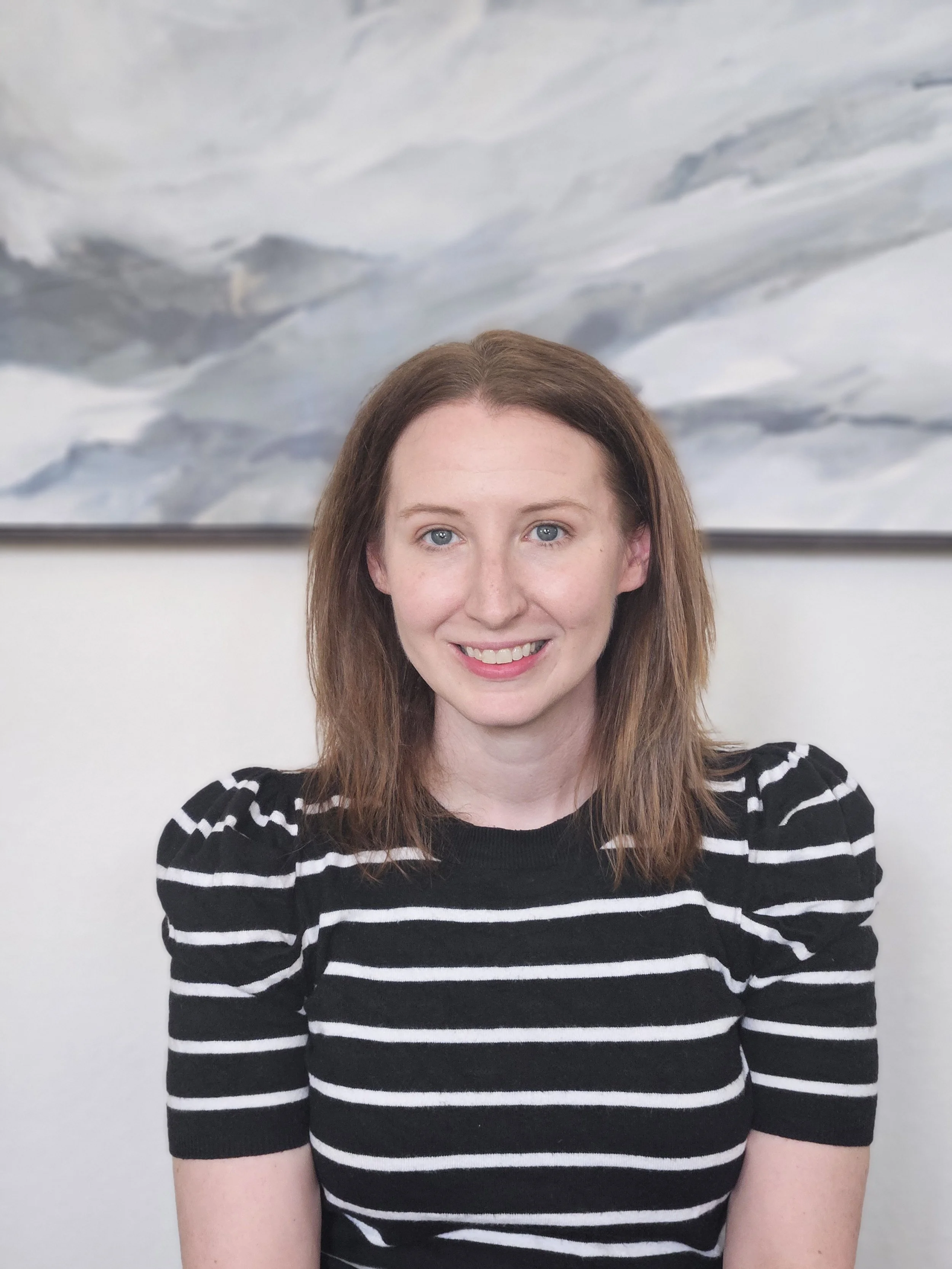 Amy Markuson with shoulder-length light brown hair and blue eyes, wearing a black and white striped top, smiling in front of an abstract mountain painting offering individual online therapy.