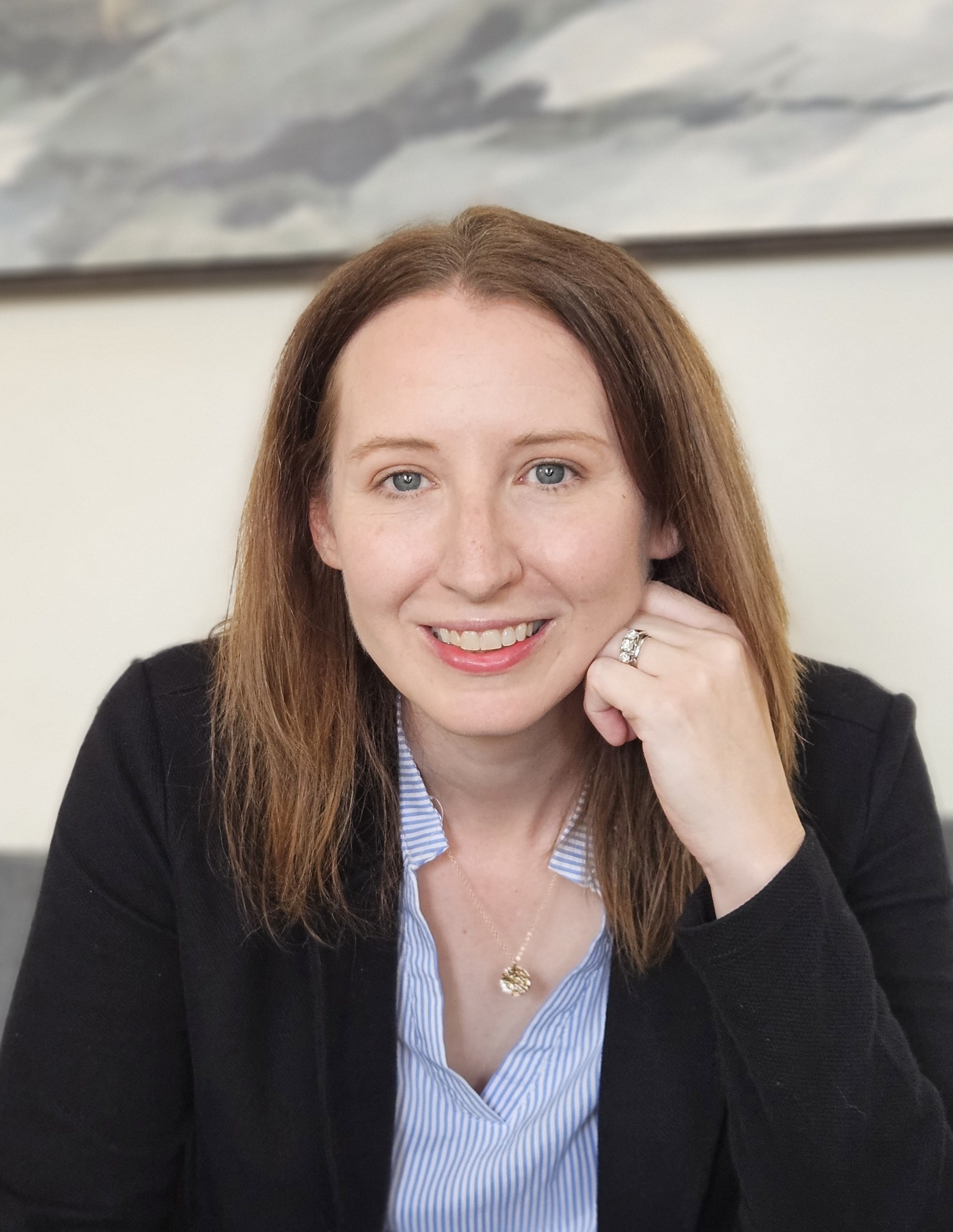 Amy Markuson with light skin, shoulder-length brown hair, and blue eyes smiling and resting her chin on her hand, wearing a black blazer, a striped blue and white shirt, a necklace, and a ring offering free consultation for virtual therapy.