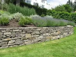 Stone retaining wall with greenery and shrubs behind it, in a landscaped yard.