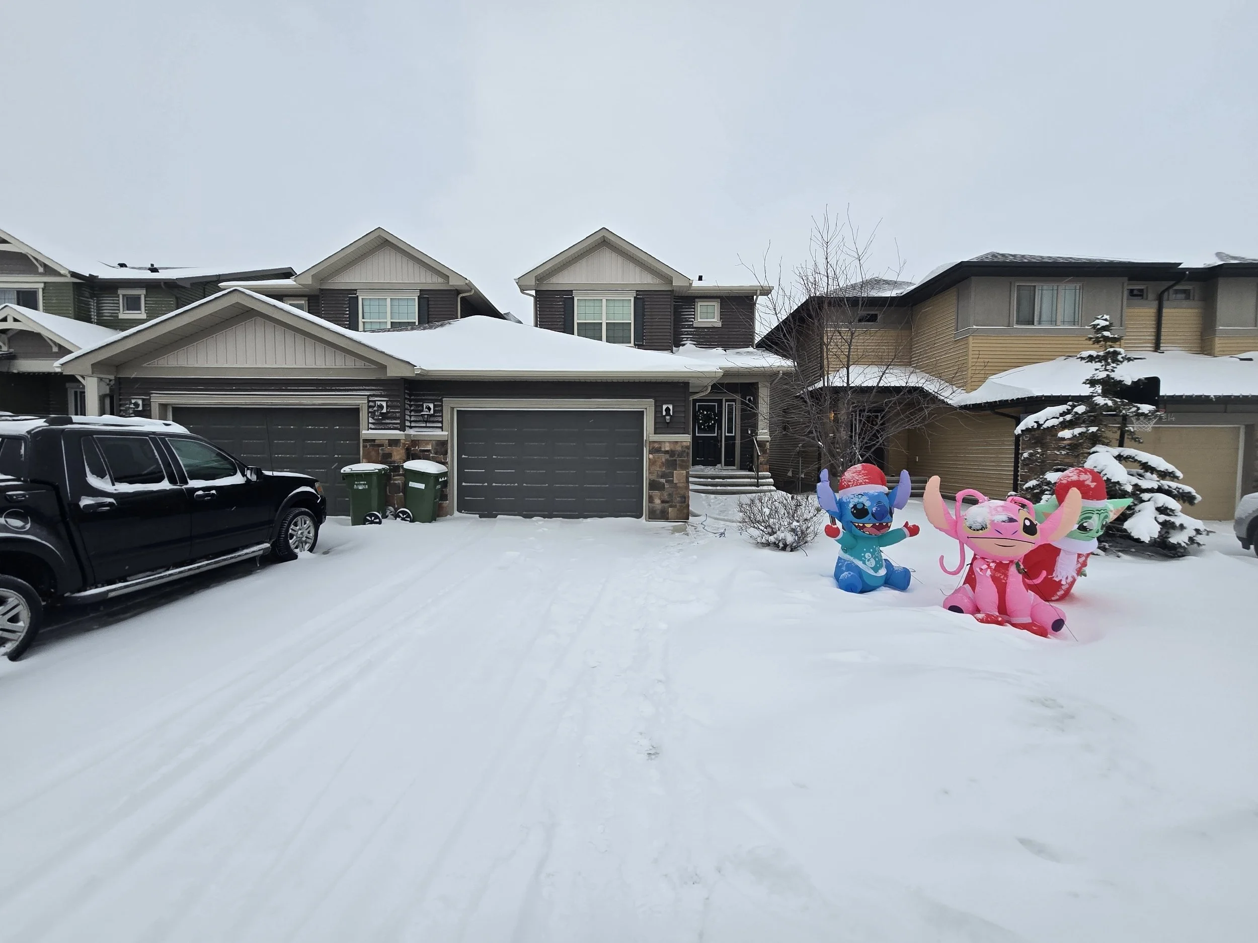 Snow-covered driveway with colorful inflatable cartoon characters in front yard, suburban houses in background, and a black truck parked on the left side.