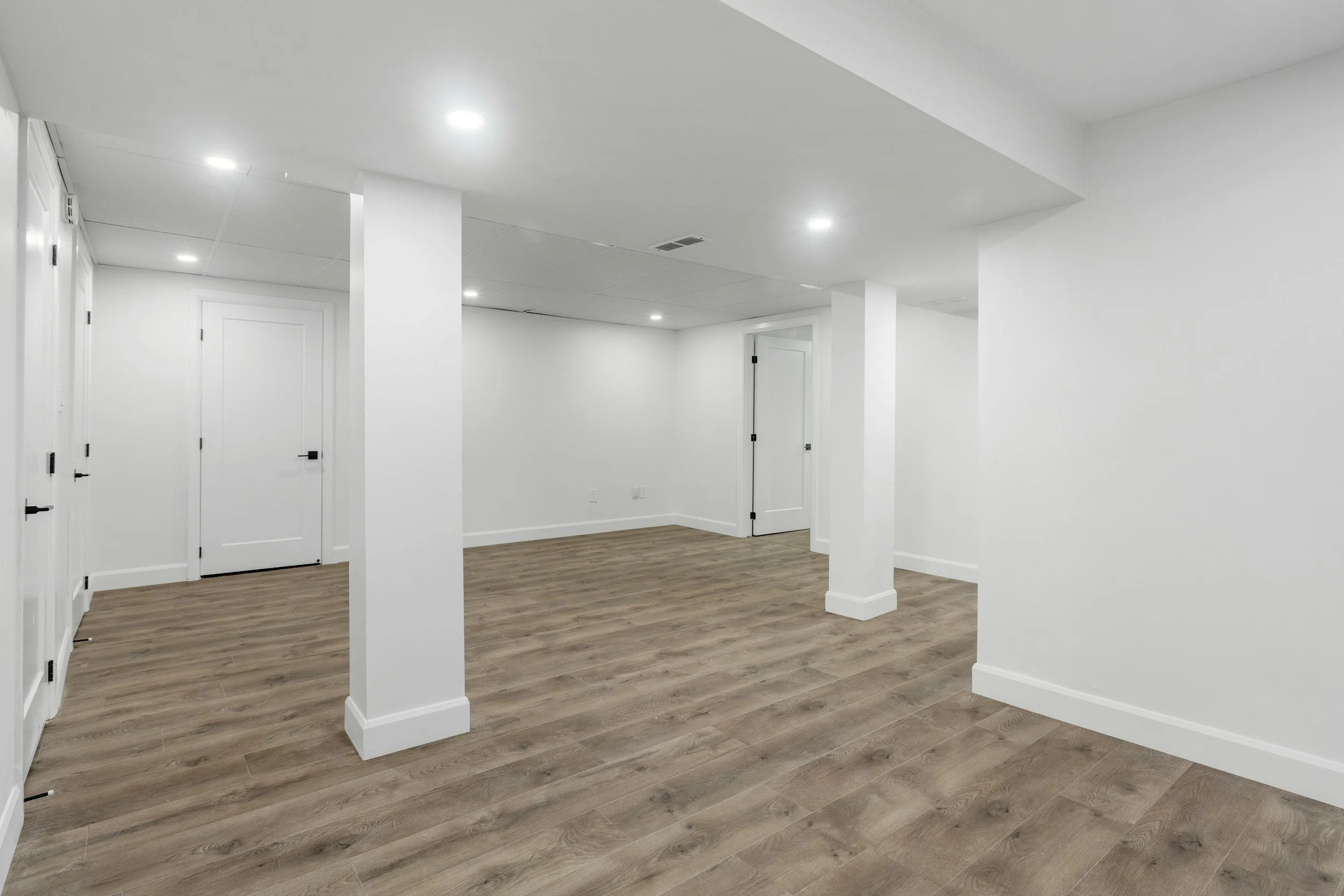 Empty white basement room with wooden floor, white walls, ceiling lights, and multiple doors.