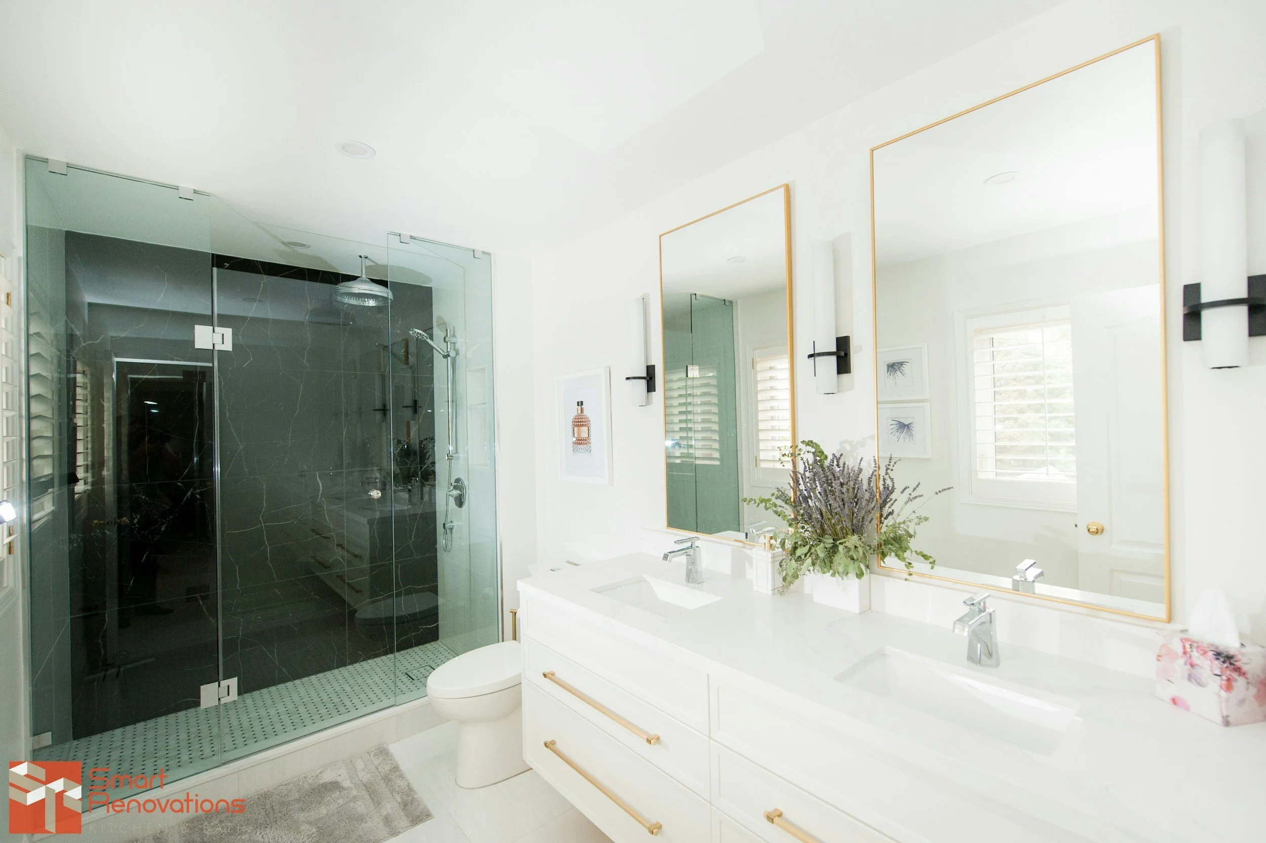 Bright modern bathroom with double sinks, large mirrors, black wall-mounted light fixtures, and a walk-in shower with black marble tiles and glass doors, decorated with a vase of lavender plants and a floral tissue box.