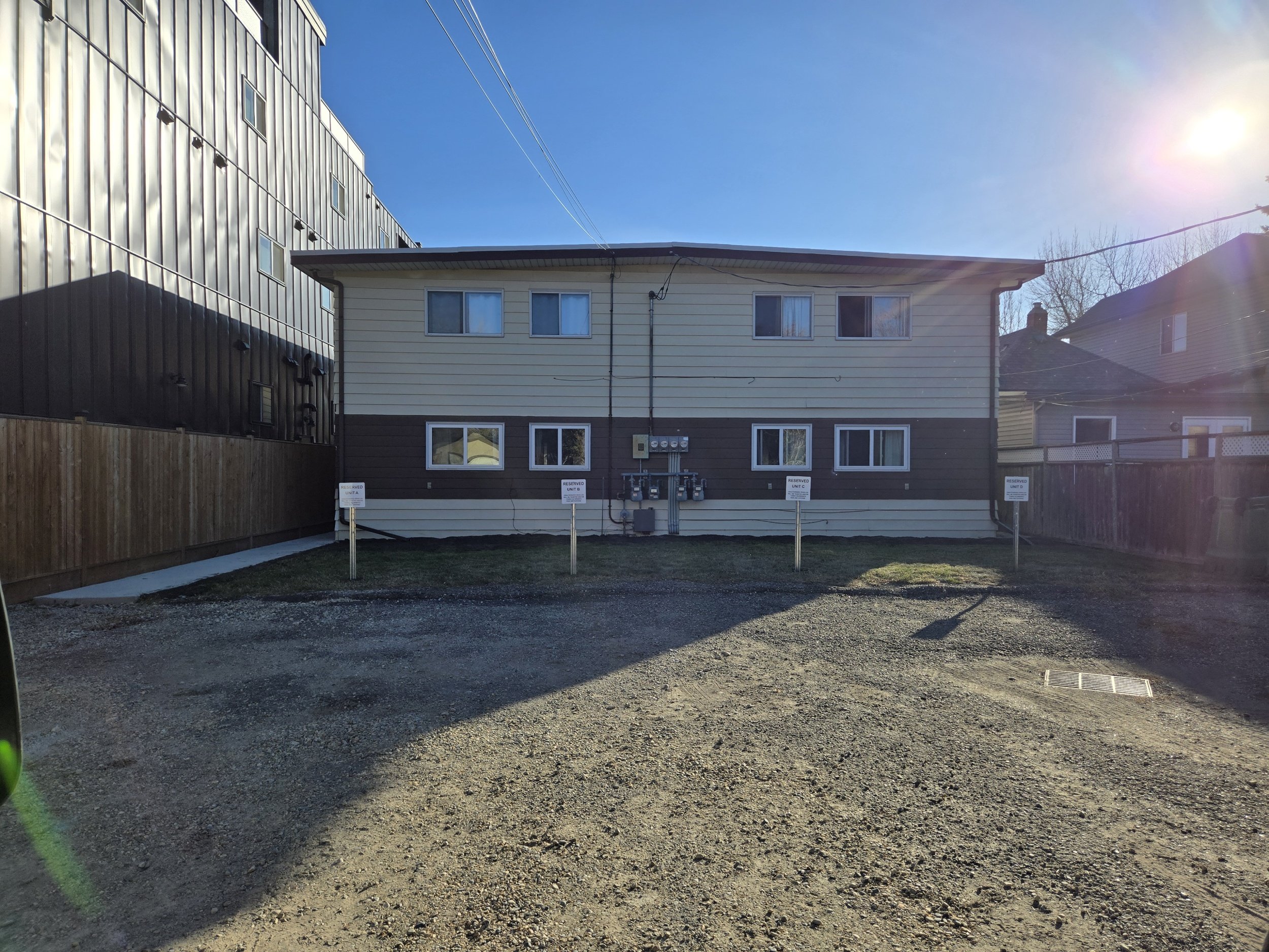 Two-story residential building with beige and brown exterior, multiple windows, and utility meters on the back wall. The building is surrounded by a gravel driveway, a grassy area, and wooden fences with a neighboring house on the right. The sky is c
