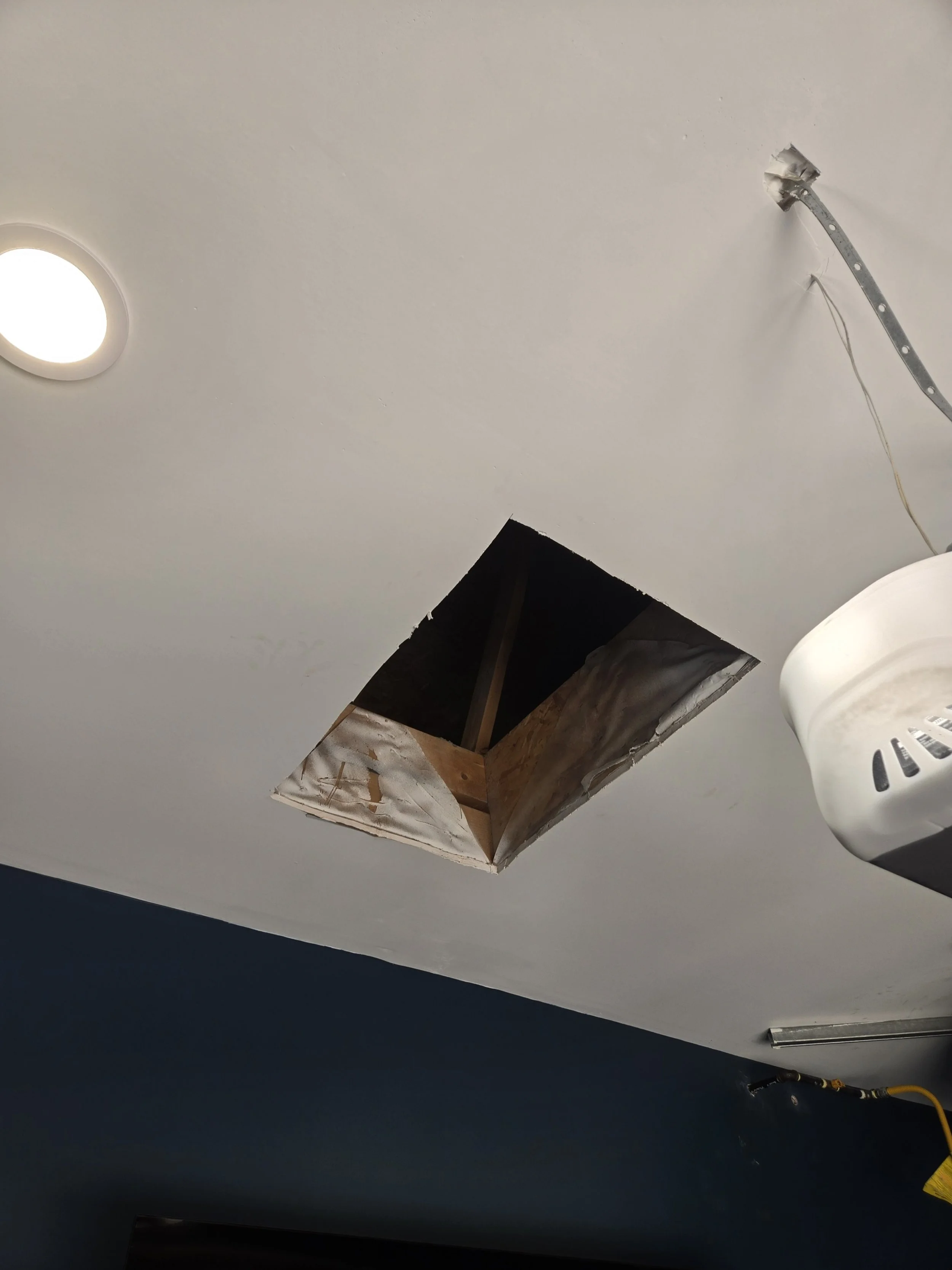 Ceiling hole exposing wooden frame in a room with a blue wall and a white ceiling light.