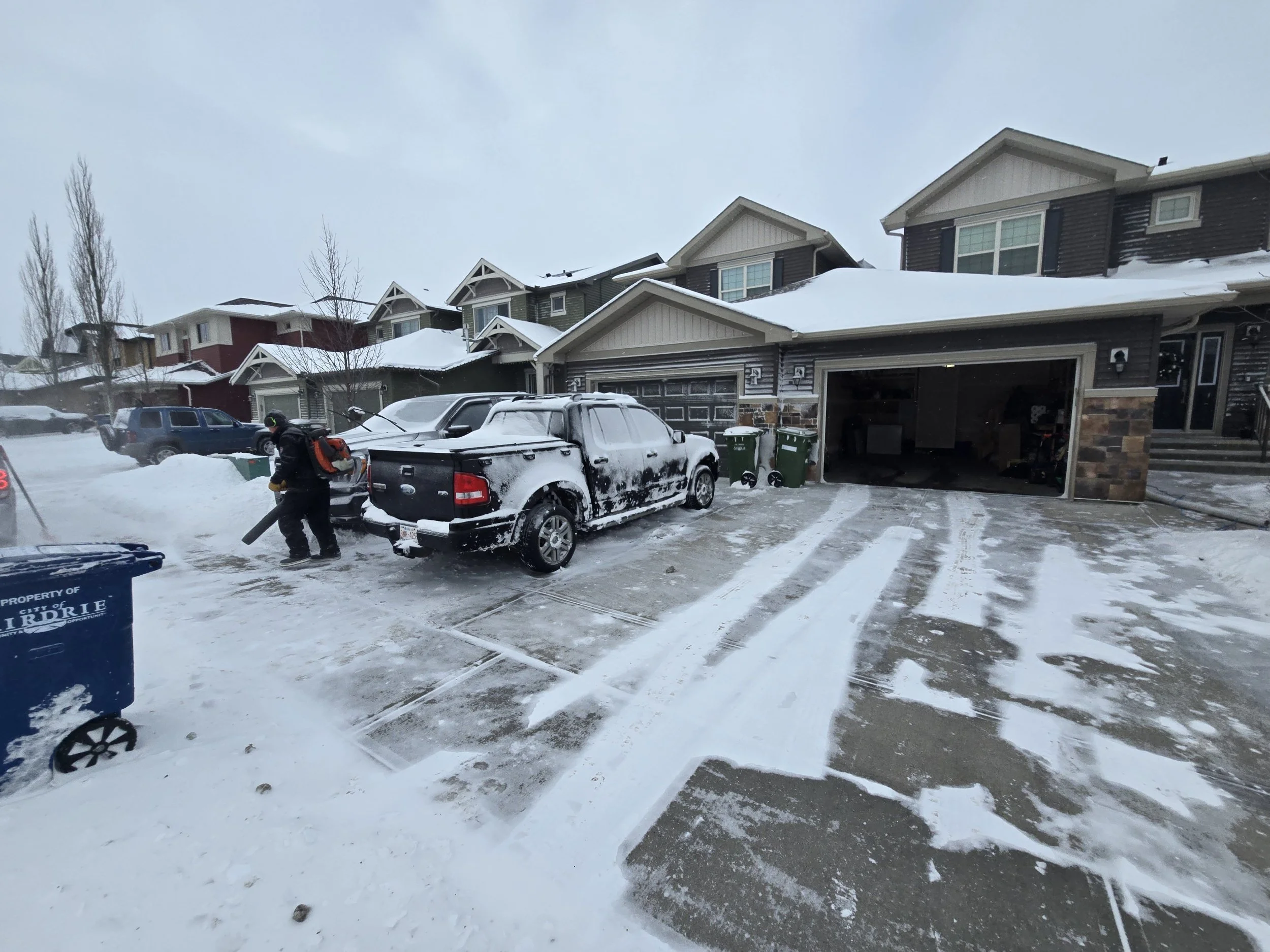 Snowy residential driveway with a person wearing winter clothing and a backpack, using a leaf blower on the snow-covered driveway. Multiple houses with garage doors and cars are visible in the background.