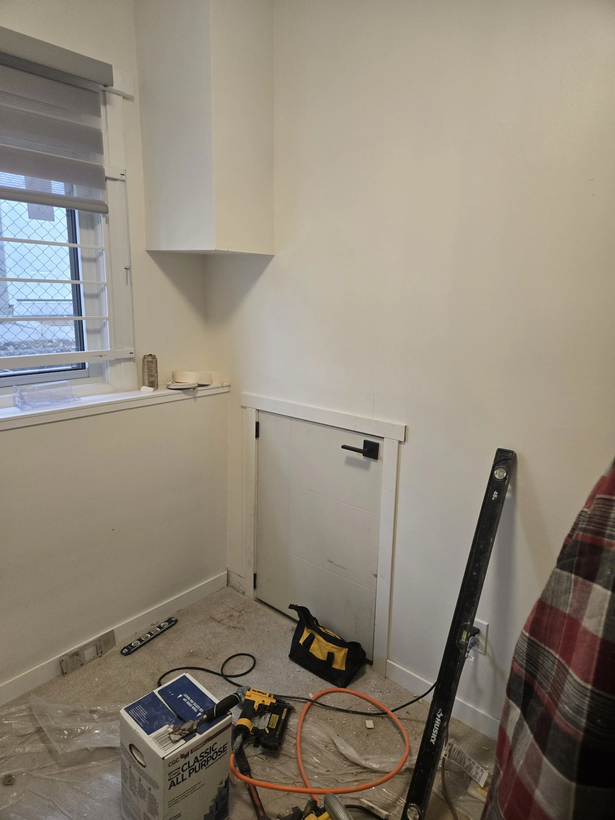 A small room with a window, a level on the floor, a toolbox, a cordless drill, an extension cord, a paint roller, a small door with a handle, and some items on the windowsill.