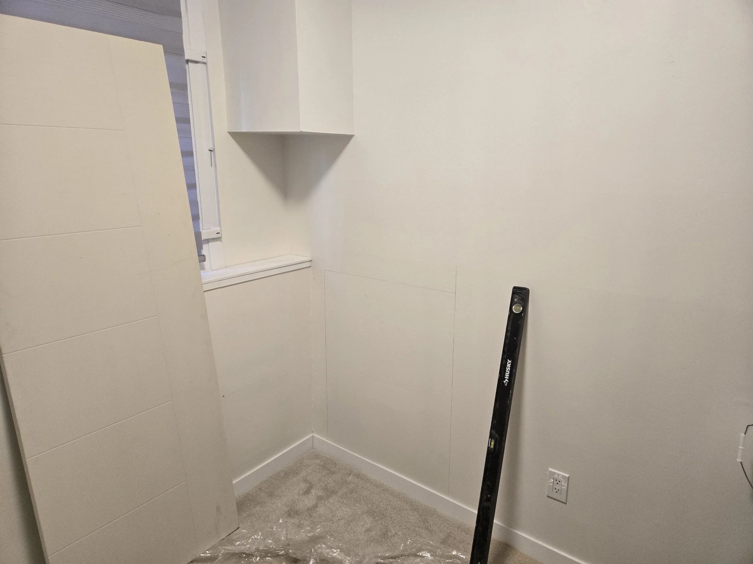 A small room with light-colored walls, a window with blinds, a level tool propped against the wall, and an electrical outlet near the floor.