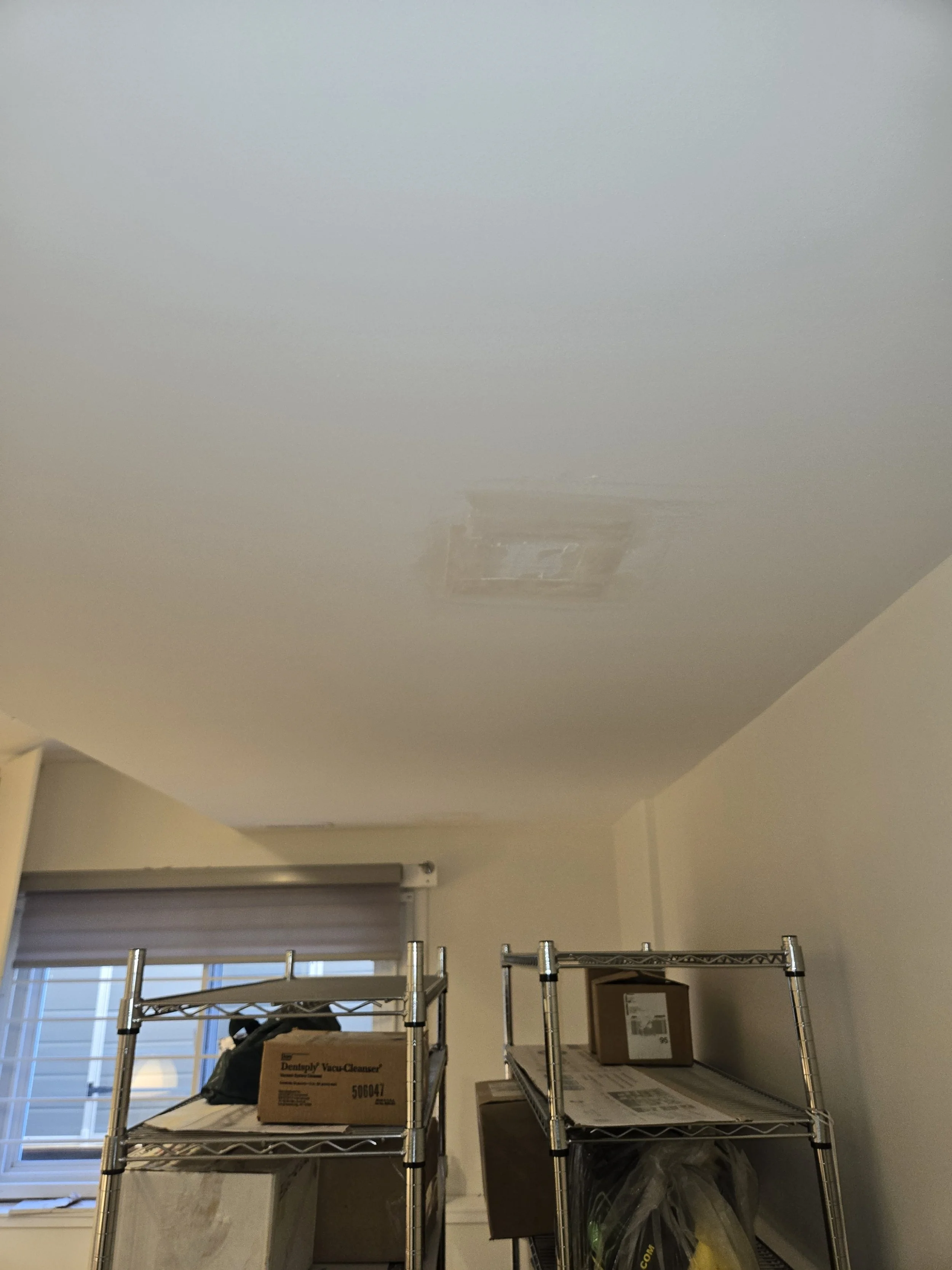 Ceiling with a patch of uneven texture or repair in a room with shelves, boxes, and a window with blinds.